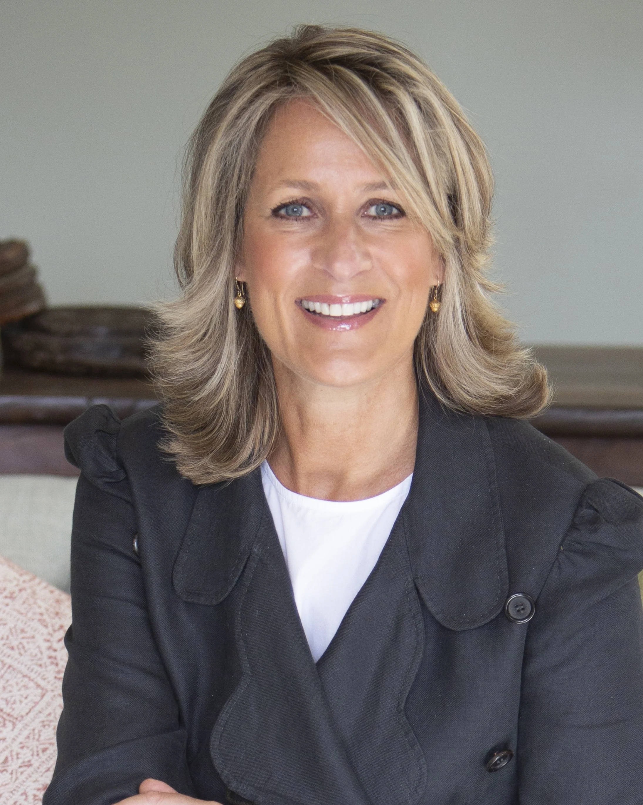 A woman with shoulder-length blonde hair, blue eyes, and a big smile, wearing a black blazer with a white top, sitting indoors with a blurred background.