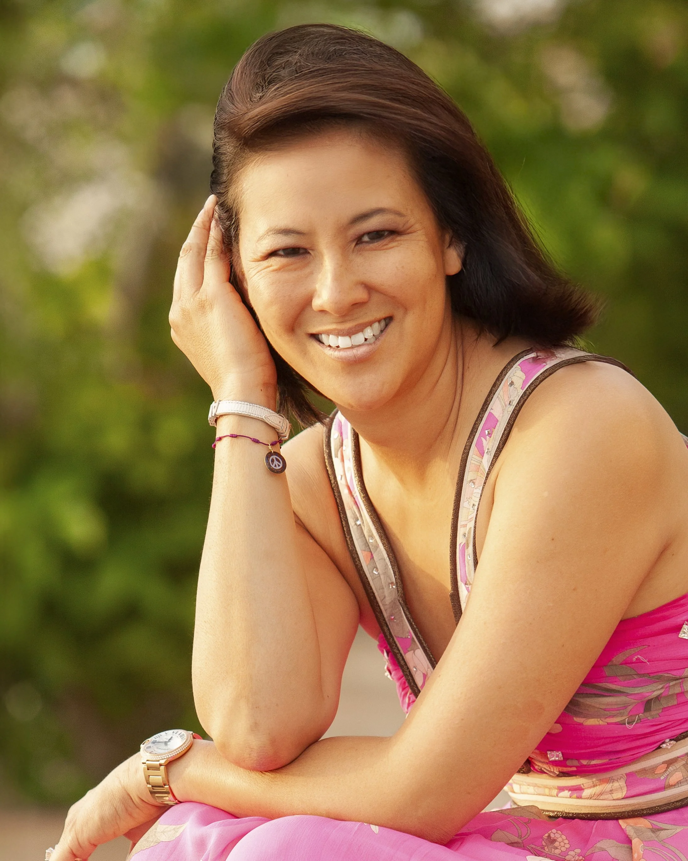 A woman with short dark hair smiling outdoors, wearing a pink and floral sleeveless top and jewelry.