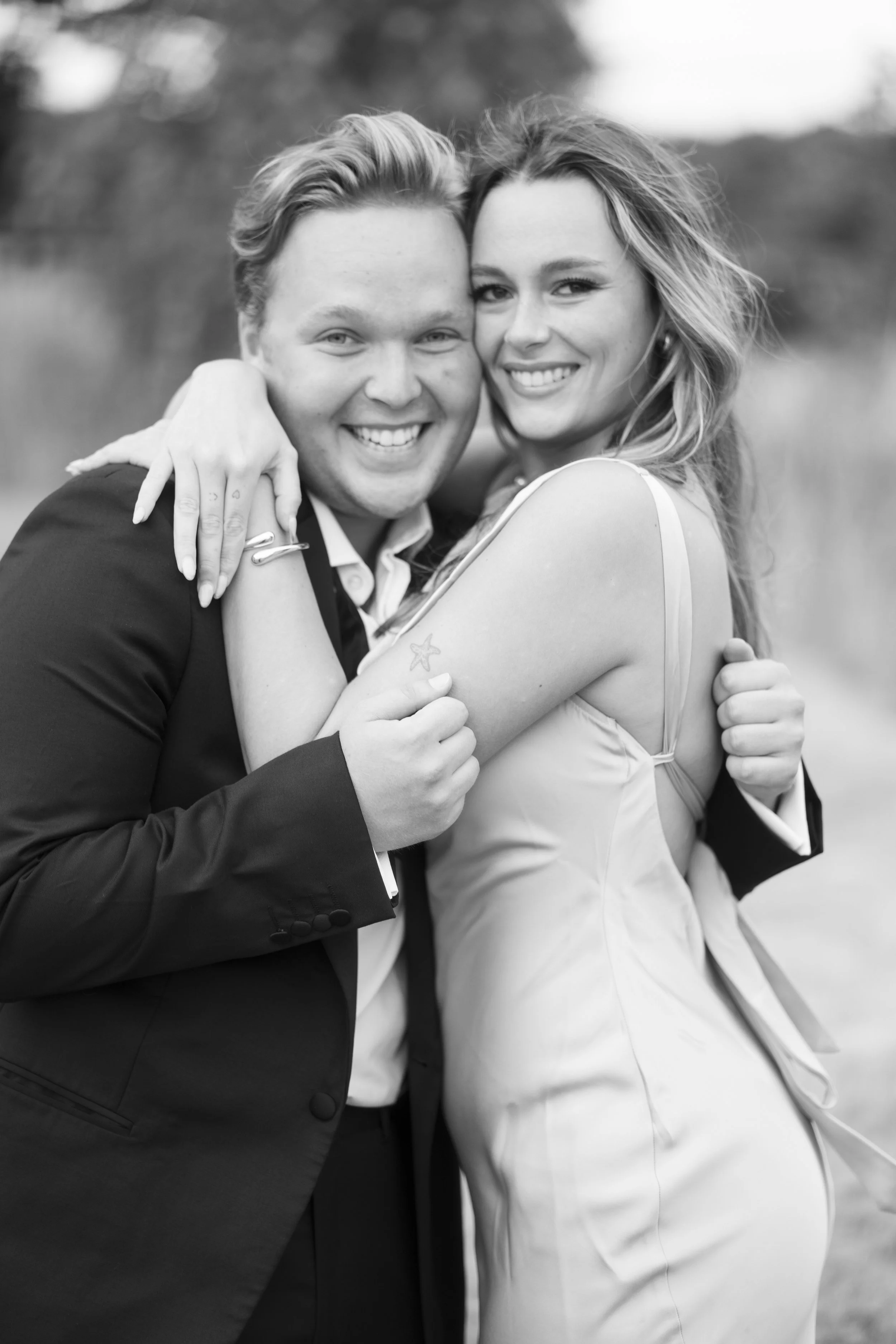 A smiling couple embracing outdoors, with the woman wearing a sleeveless dress and the man in a suit, both looking happy.