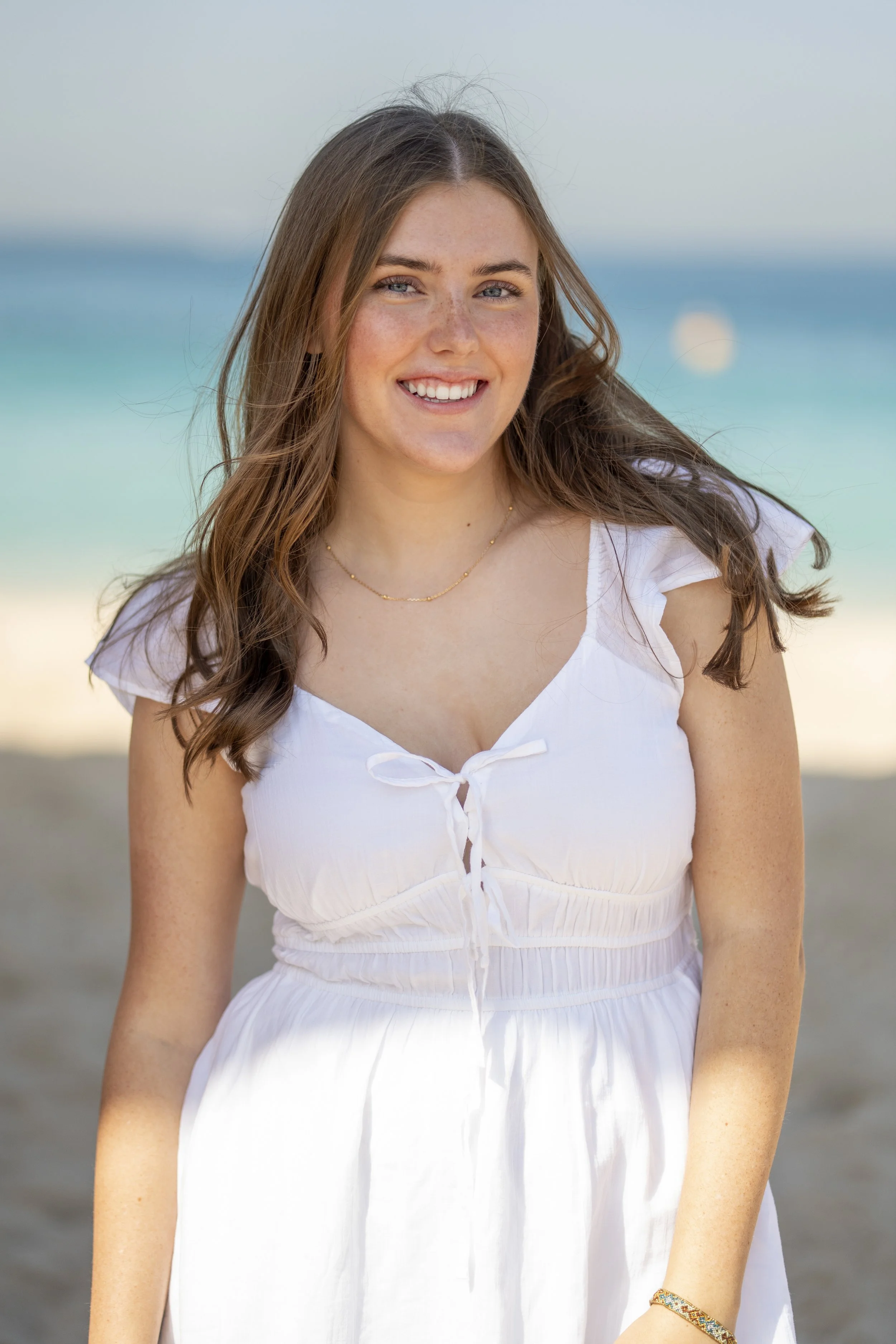 A young woman with long brown hair, wearing a white dress, smiling on a beach with ocean waves in the background.