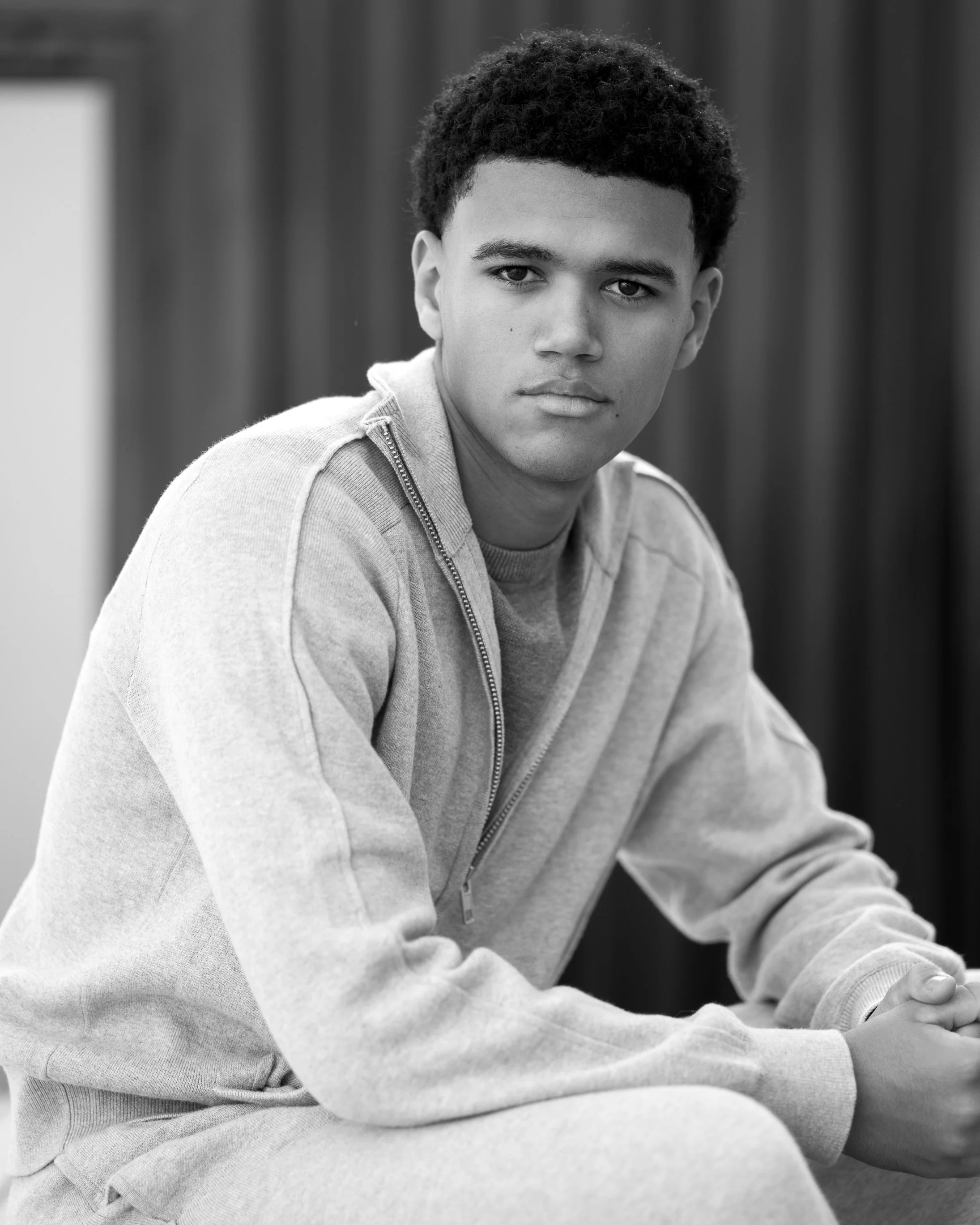 A young man with short curly hair sitting indoors, looking directly at the camera, wearing a light-colored zip-up sweatshirt.