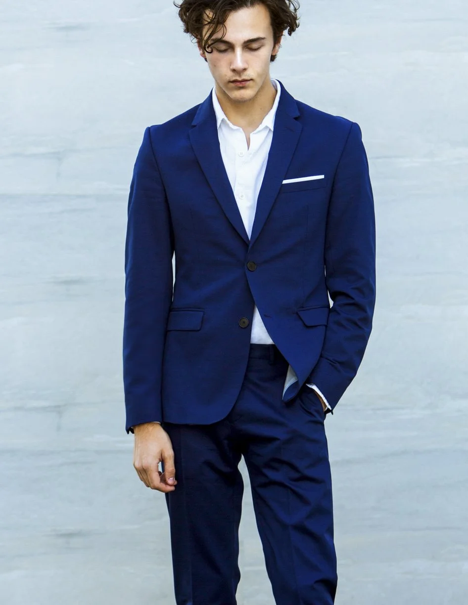 Young man in a navy blue suit with a white shirt, standing with his eyes closed against a light background.