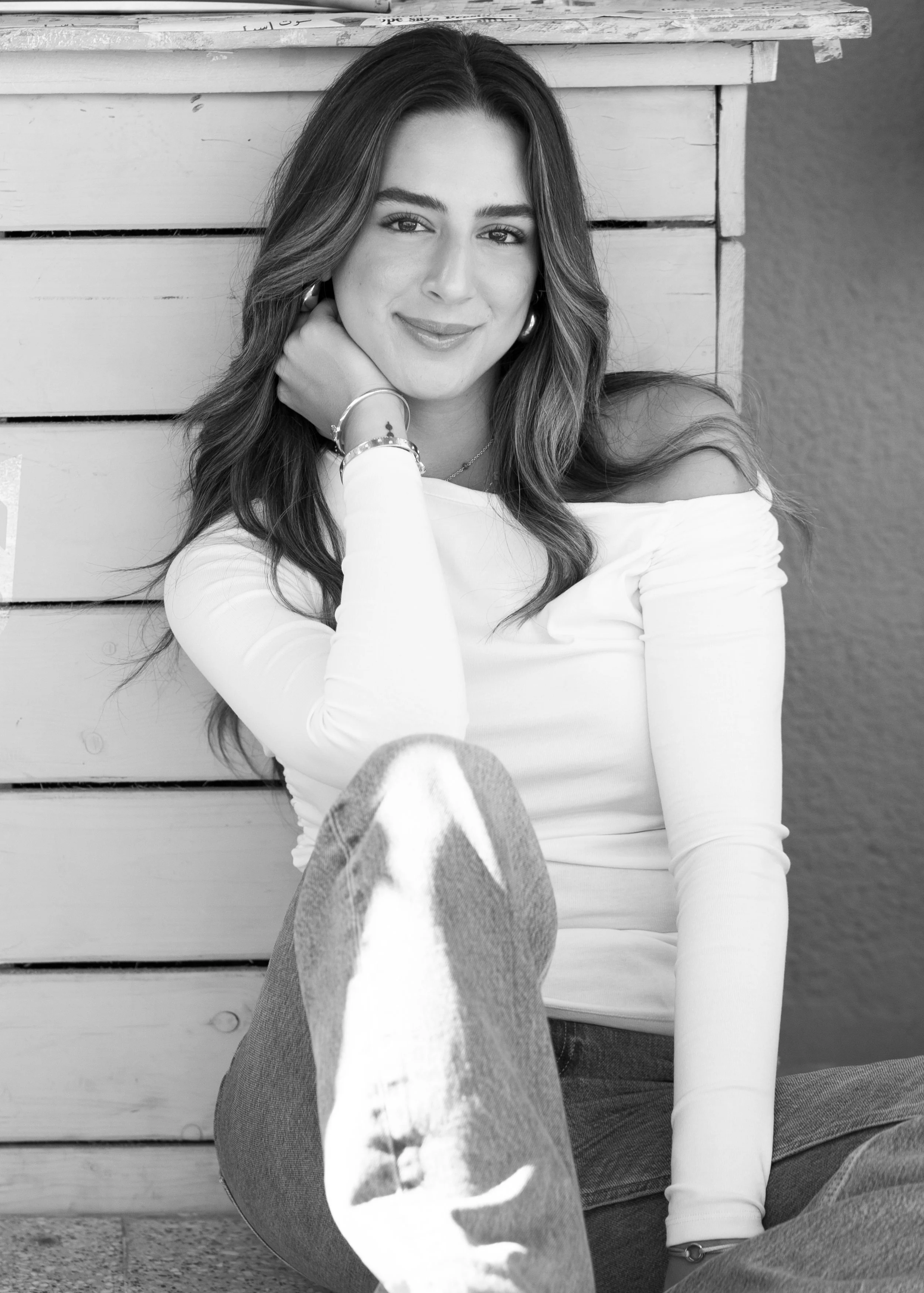 Black and white photo of a young woman with long wavy hair, wearing an off-shoulder white top and jeans, sitting and leaning against a wooden wall with a smile.