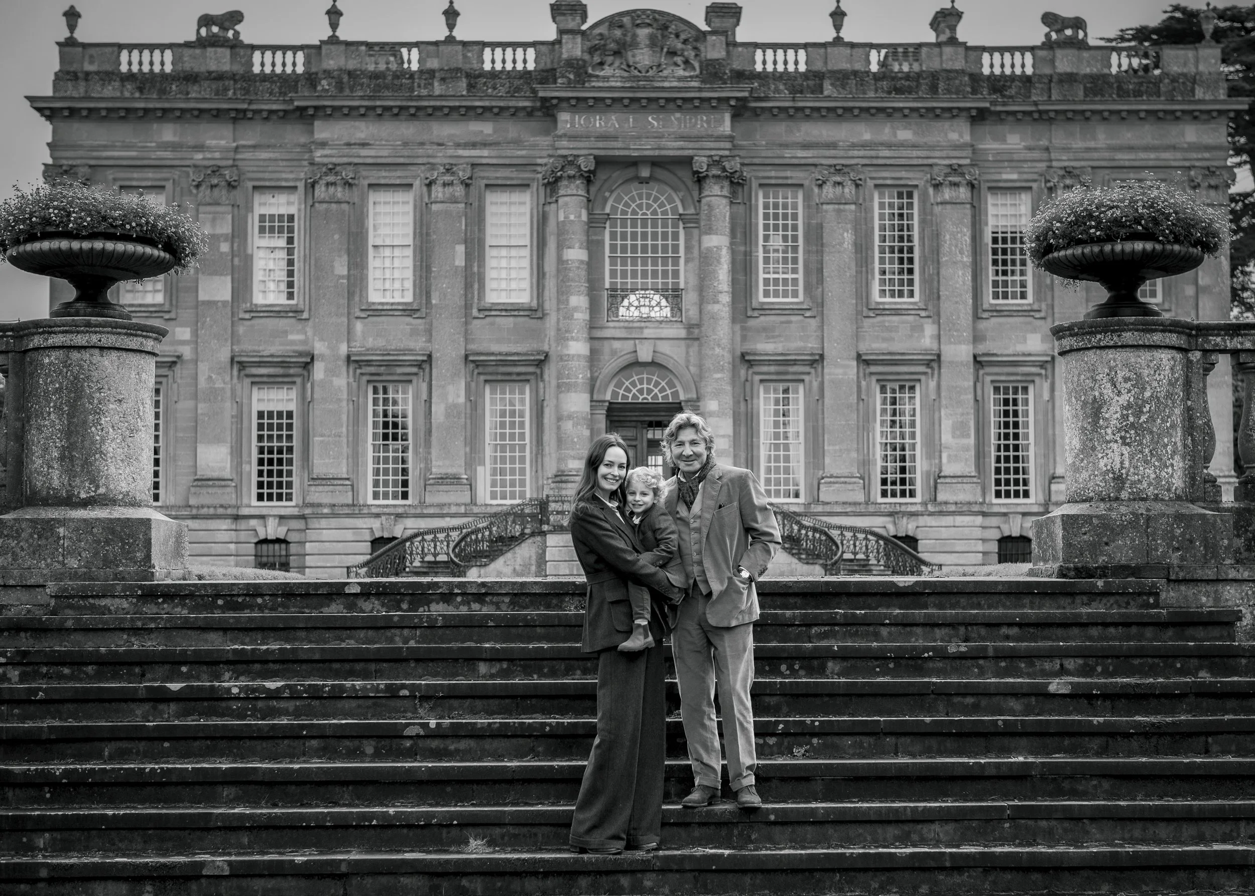 A family of three standing on stone steps in front of a grand historic building with large windows and ornate architectural details, black and white photo.