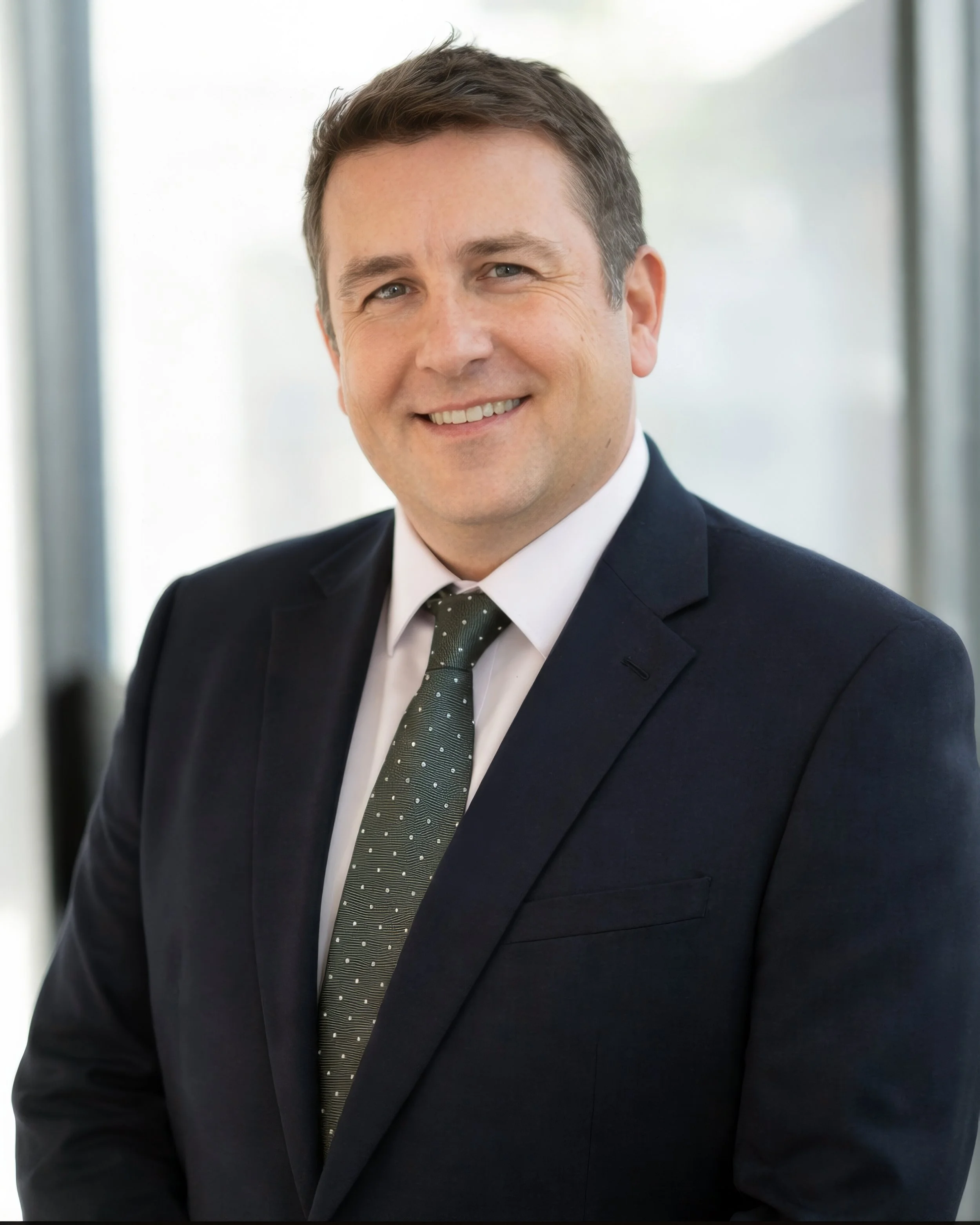 Professional headshot of a smiling man in a dark suit, white shirt, and dotted tie, with a blurred office background.
