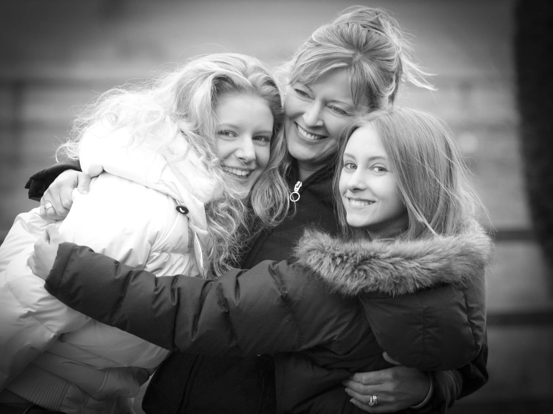 Three women embracing and smiling outdoors, dressed in winter jackets.
