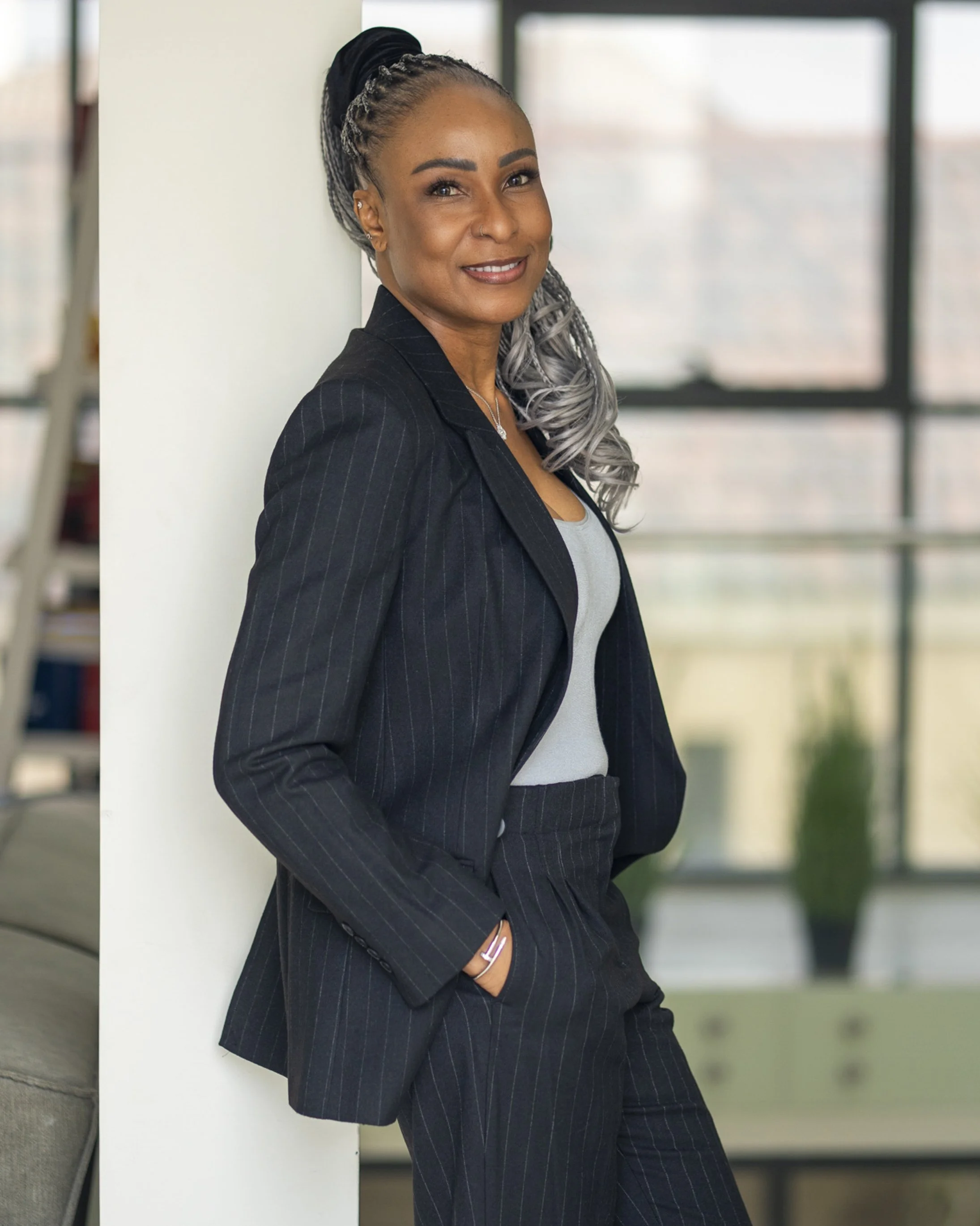 A professional woman with braided hair styled in a gray and black ombre, wearing a black pinstripe suit with a gray top, standing and smiling in a modern office environment with large windows.