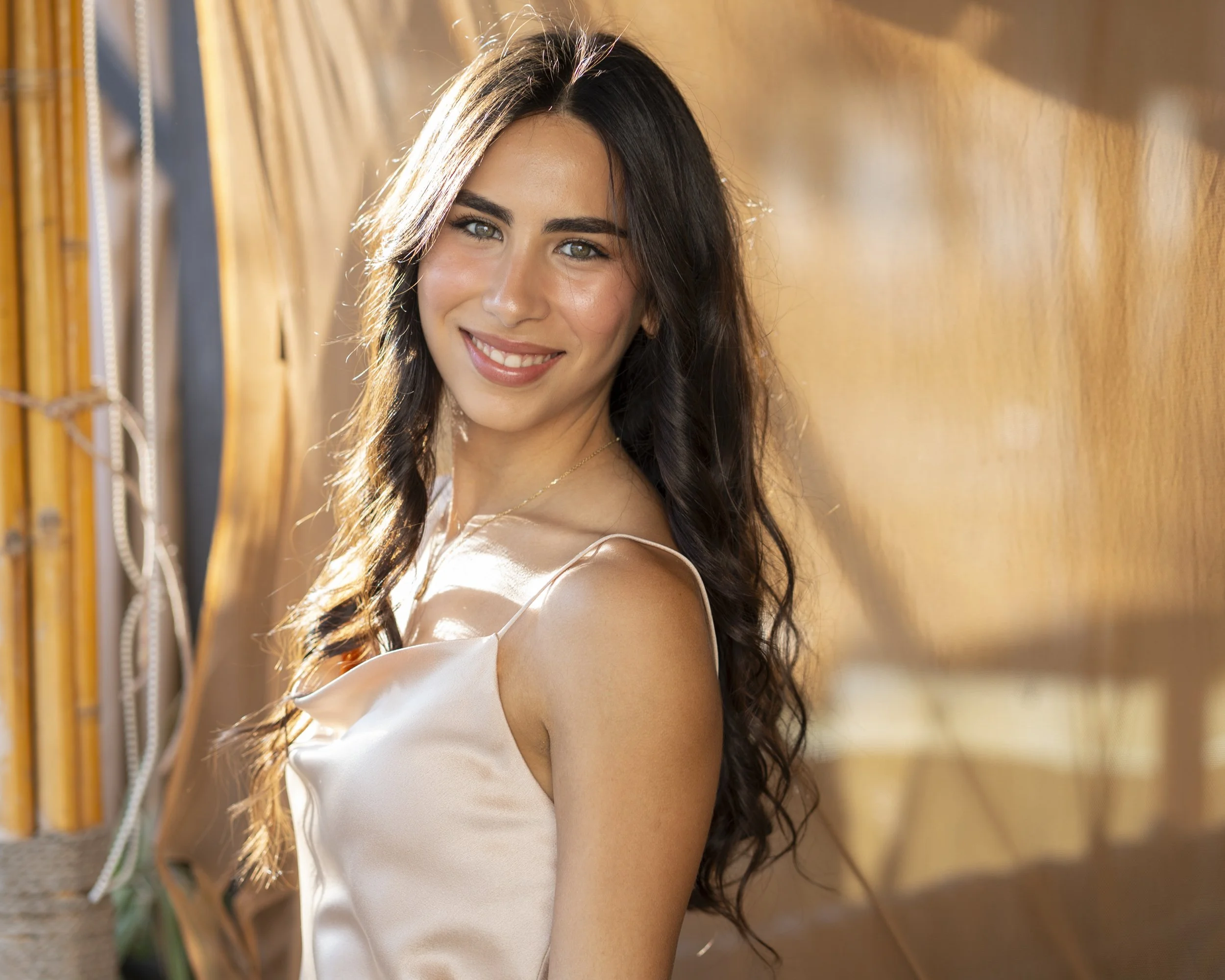A young woman with long, wavy brown hair and blue eyes, wearing a satin camisole top, smiling at the camera, with sunlight illuminating her face and a wooden background.