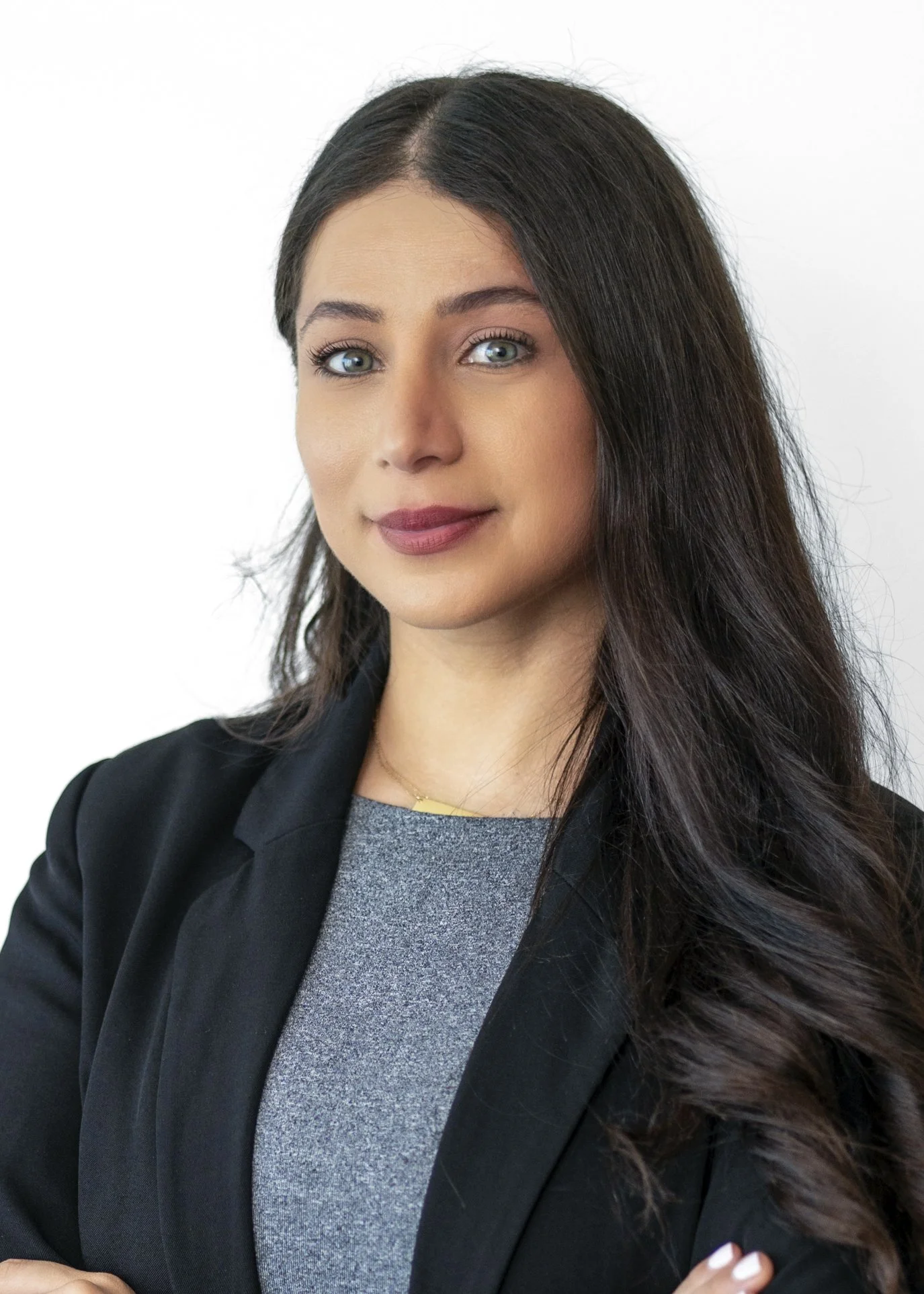 A woman with long dark hair, blue eyes, wearing a black blazer over a gray top, standing against a white background.