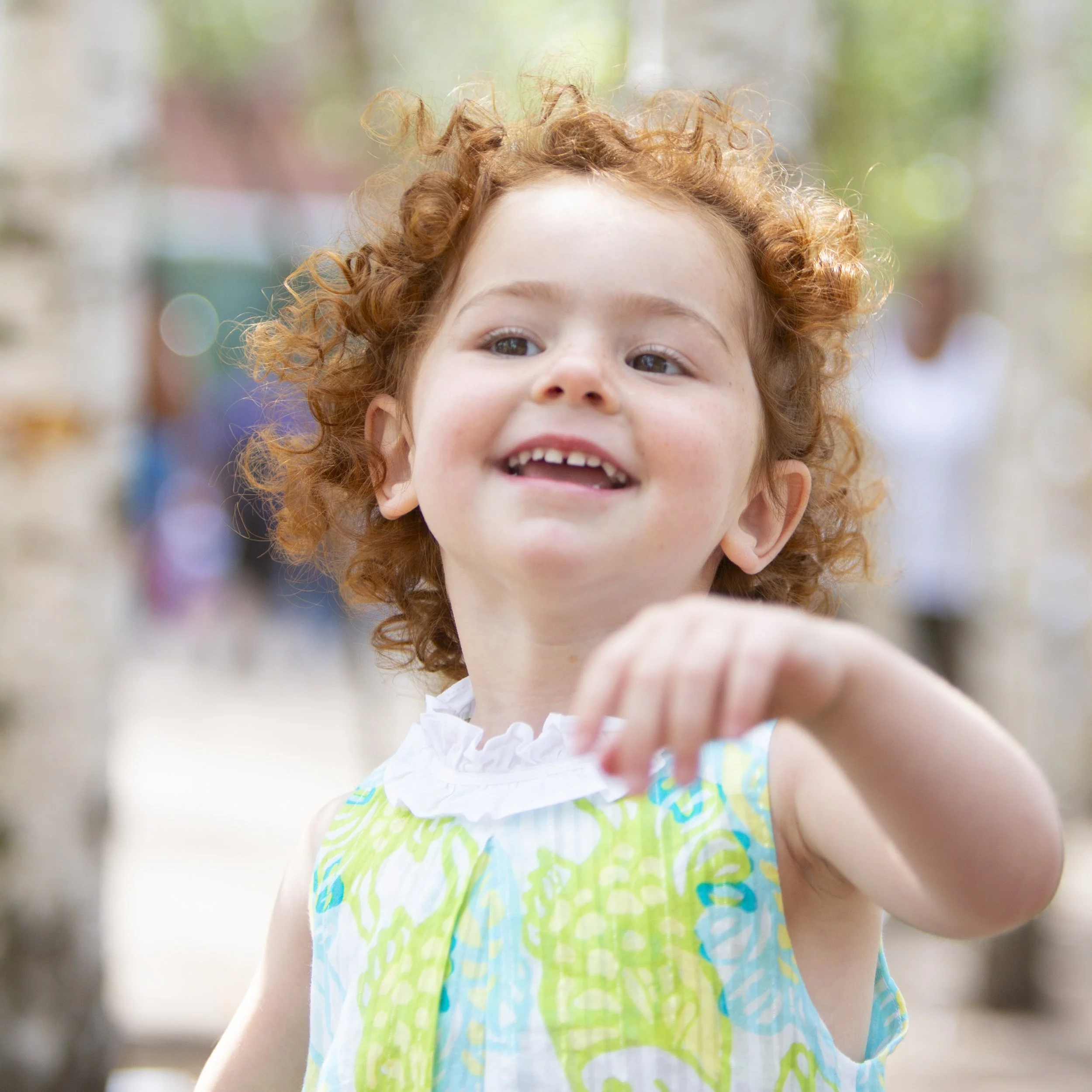 A young girl with curly red hair smiling and reaching out outdoors, with out-of-focus people and trees in the background.