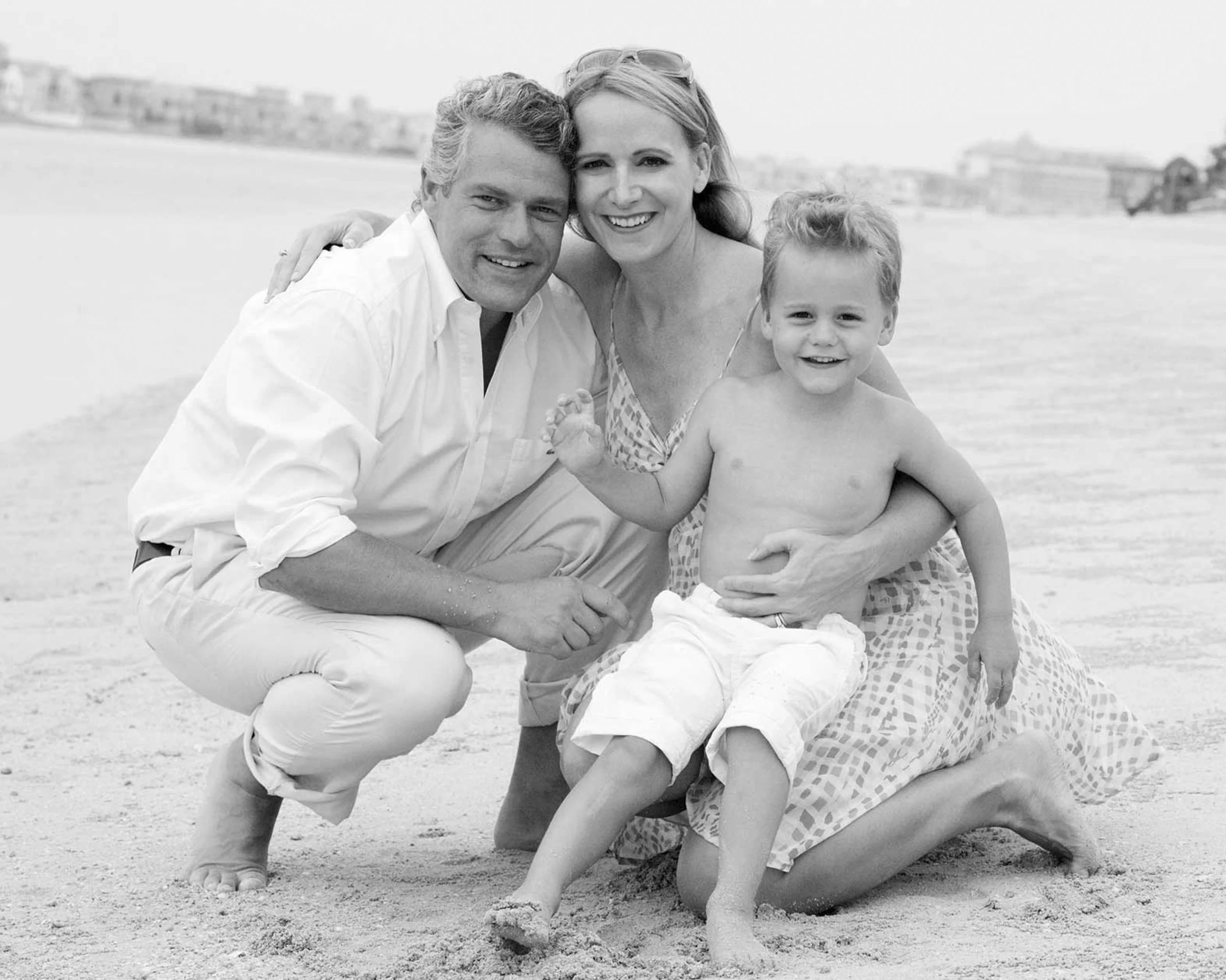 A family of three at the beach, with the mother and father kneeling and the young boy standing and smiling, all together.