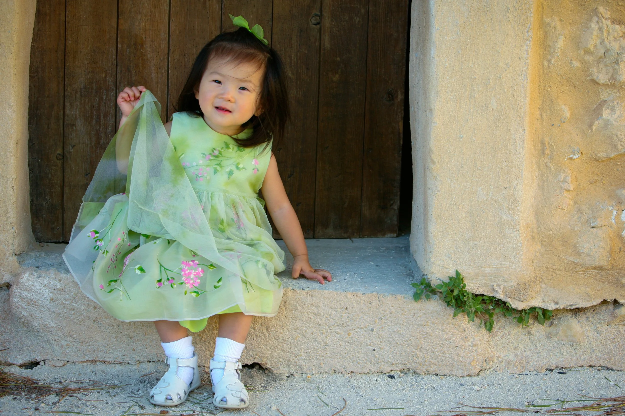 A young girl in a green floral dress sitting on a ledge outside near a wooden door and a stone wall, holding part of her dress.