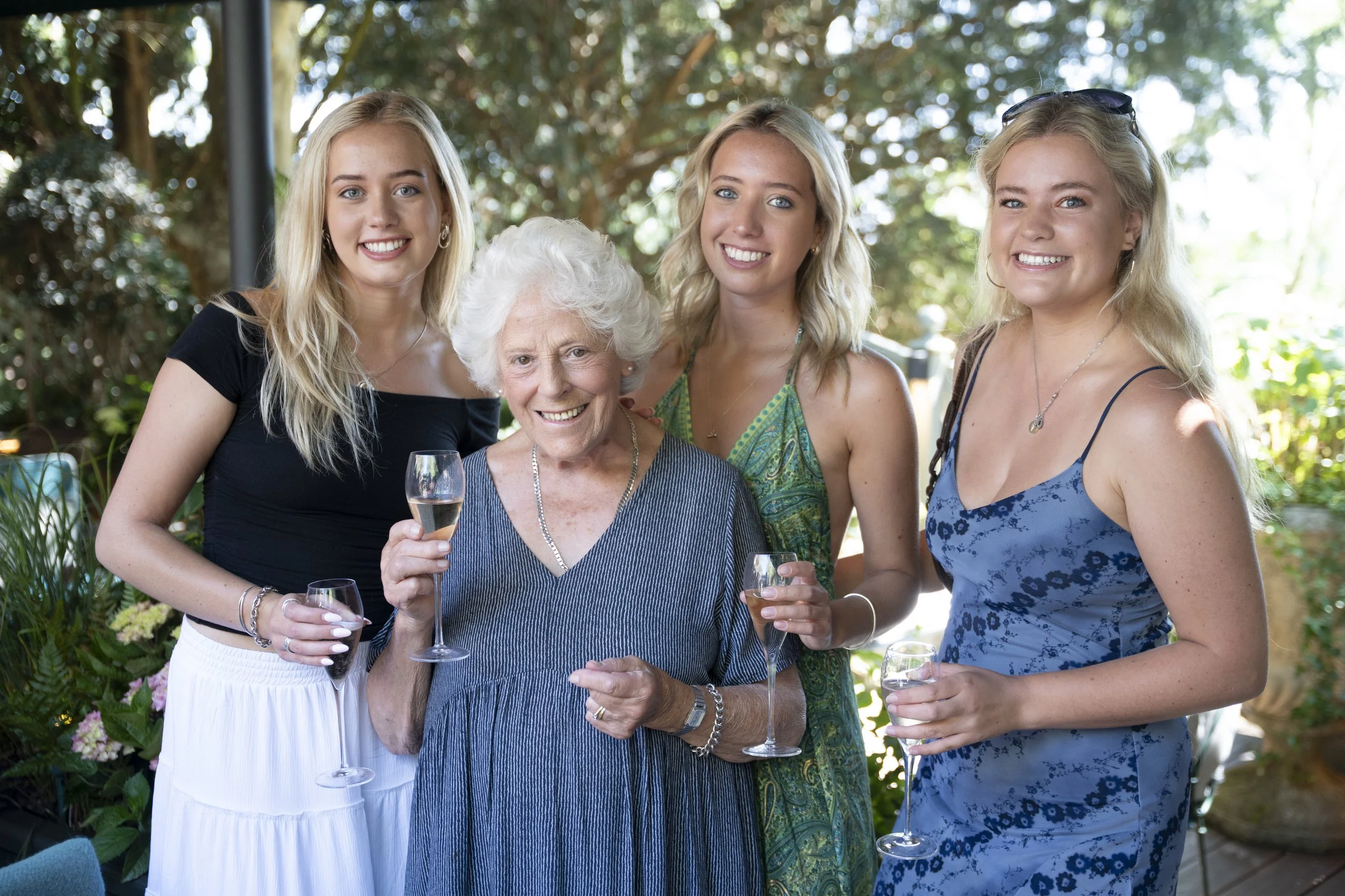 Four women, including an elderly woman in a blue dress and three younger women in casual summer dresses, smiling and holding glasses of champagne at an outdoor gathering with trees and plants in the background.
