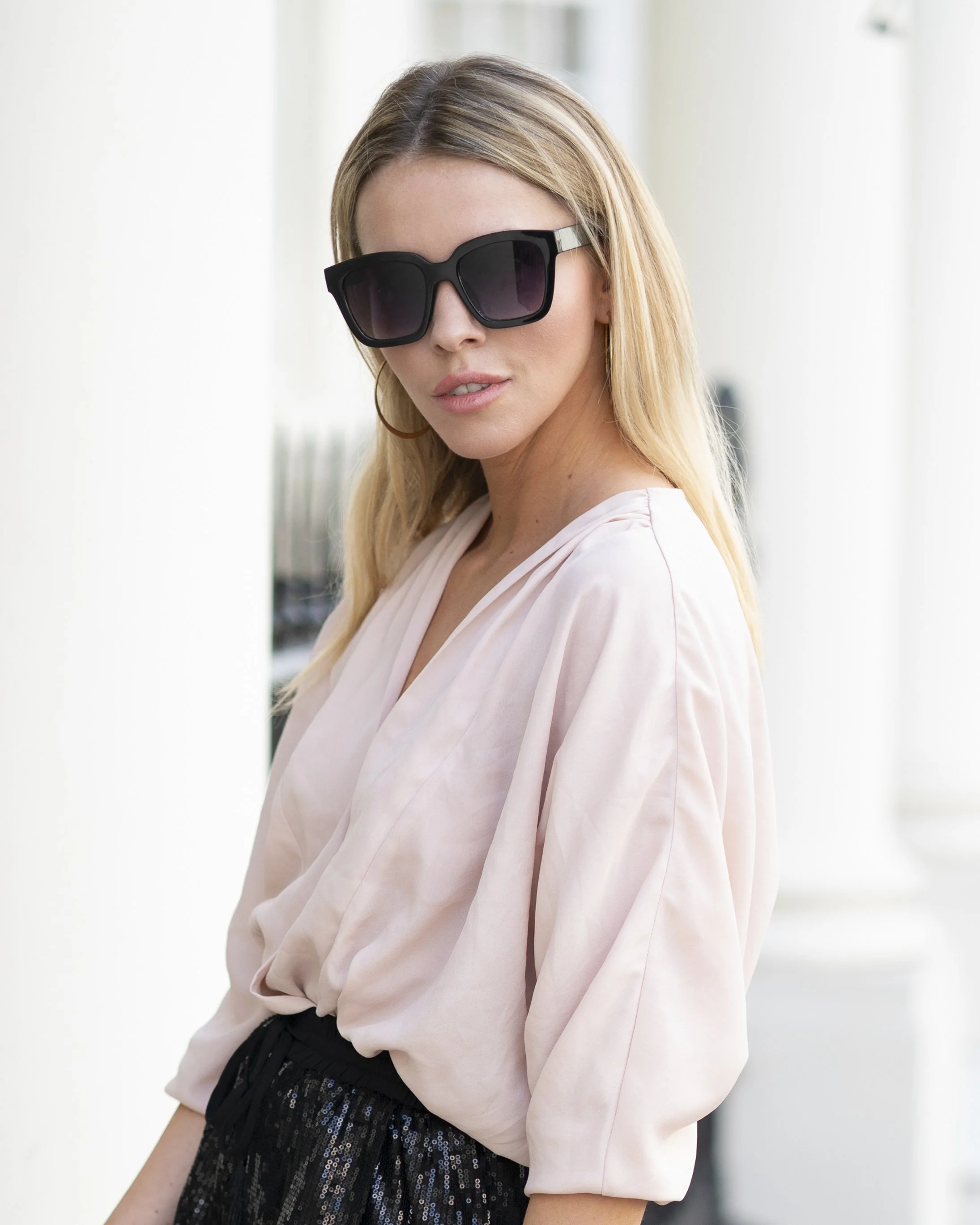 A woman with blonde hair wearing large black sunglasses, a light pink blouse, and a black sequined skirt.