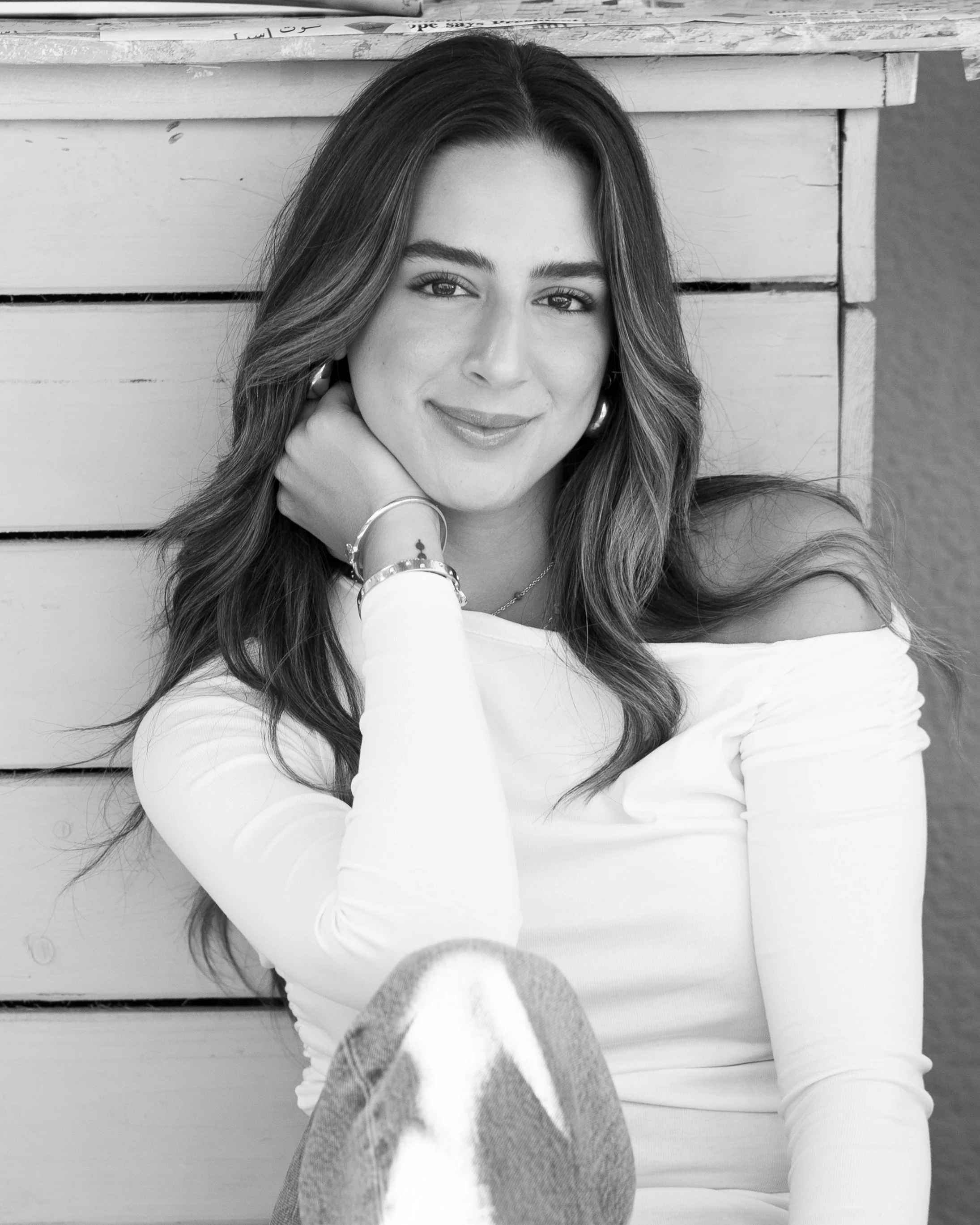 A young woman with long wavy hair styled in loose curls, smiling and sitting against a wooden wall, wearing a long sleeve top and jeans, with her hand resting near her neck.