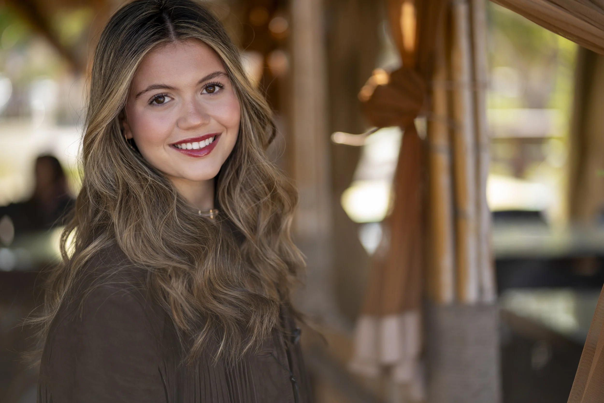 A young woman with long, wavy light brown hair smiling in an outdoor setting with wooden structures and curtains in the background.