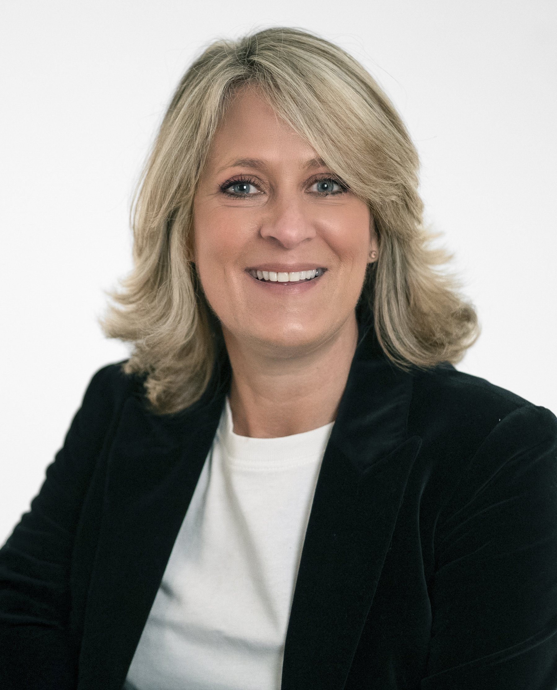 A woman with shoulder-length blonde hair, smiling, wearing a black blazer over a white top, against a plain white background.