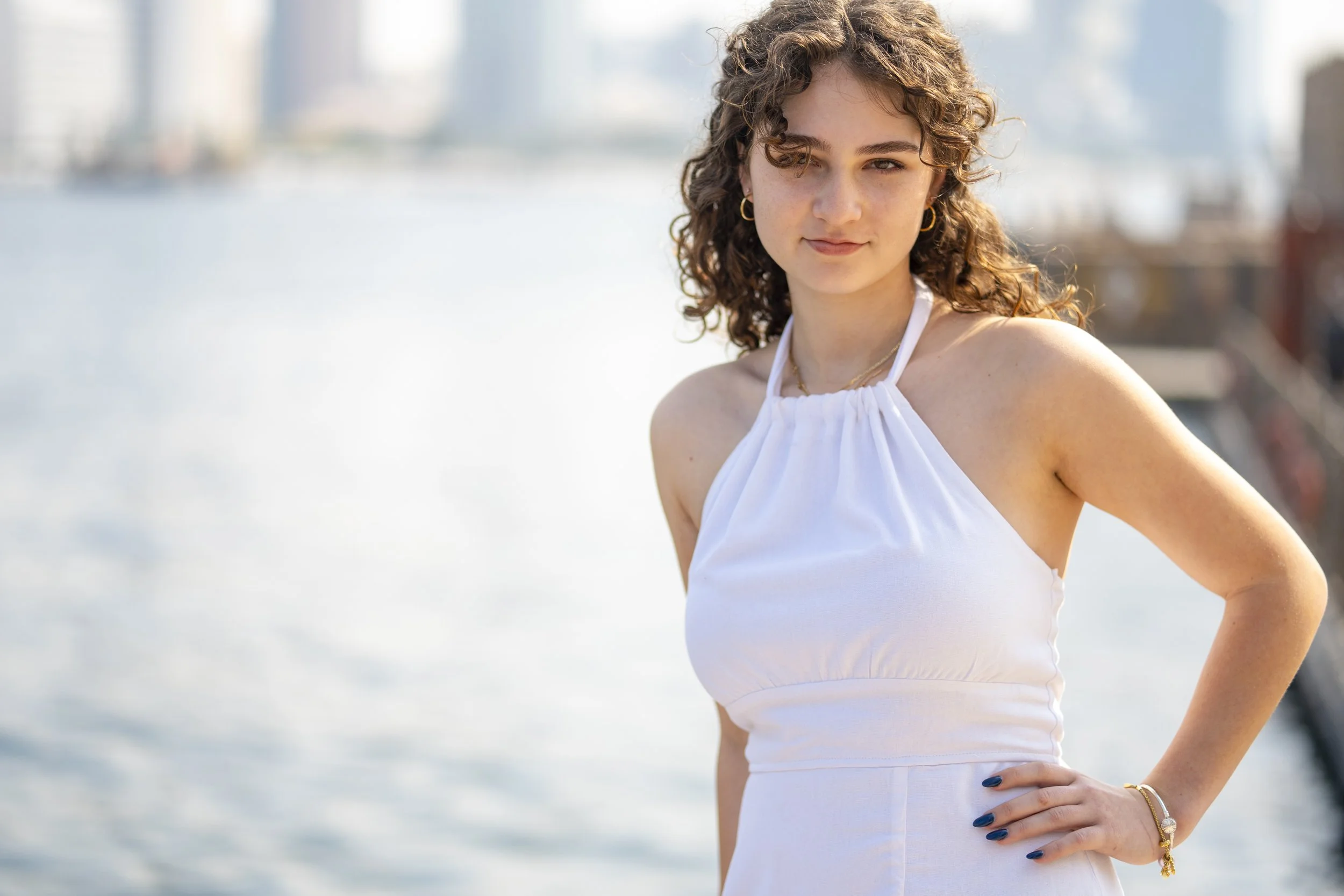 A woman with curly hair wearing a white halter dress, standing outdoors near water with a city skyline in the background.