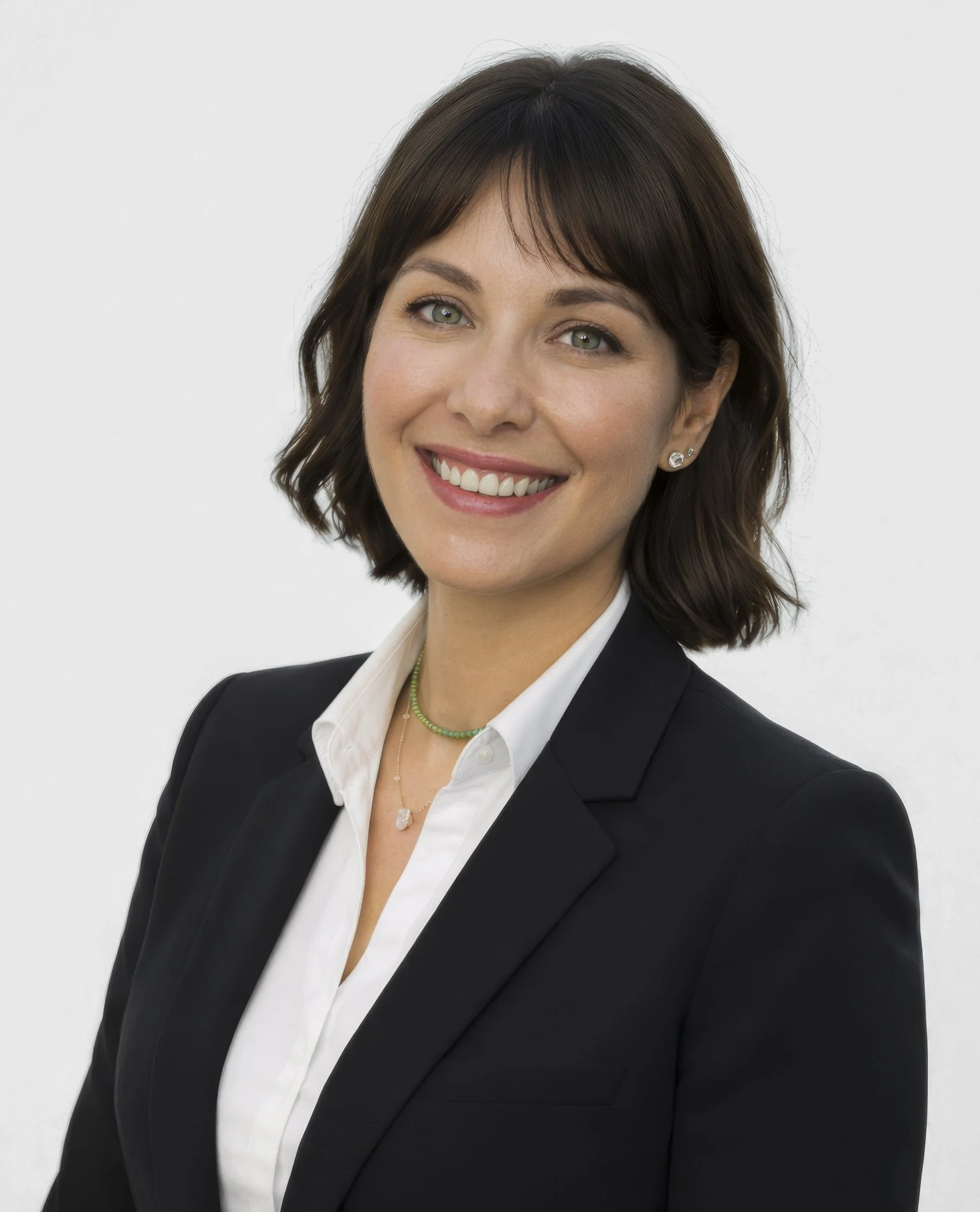 A woman with short brown hair and blue eyes smiling, wearing a black blazer, white shirt, and jewelry, standing against a white background.