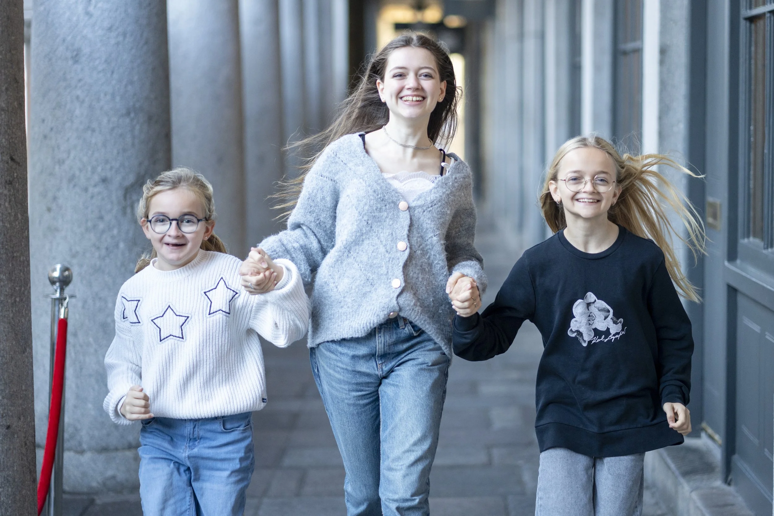 Three smiling girls holding hands and walking down a city sidewalk, with one girl in the middle and two younger girls on her sides, all dressed casually.