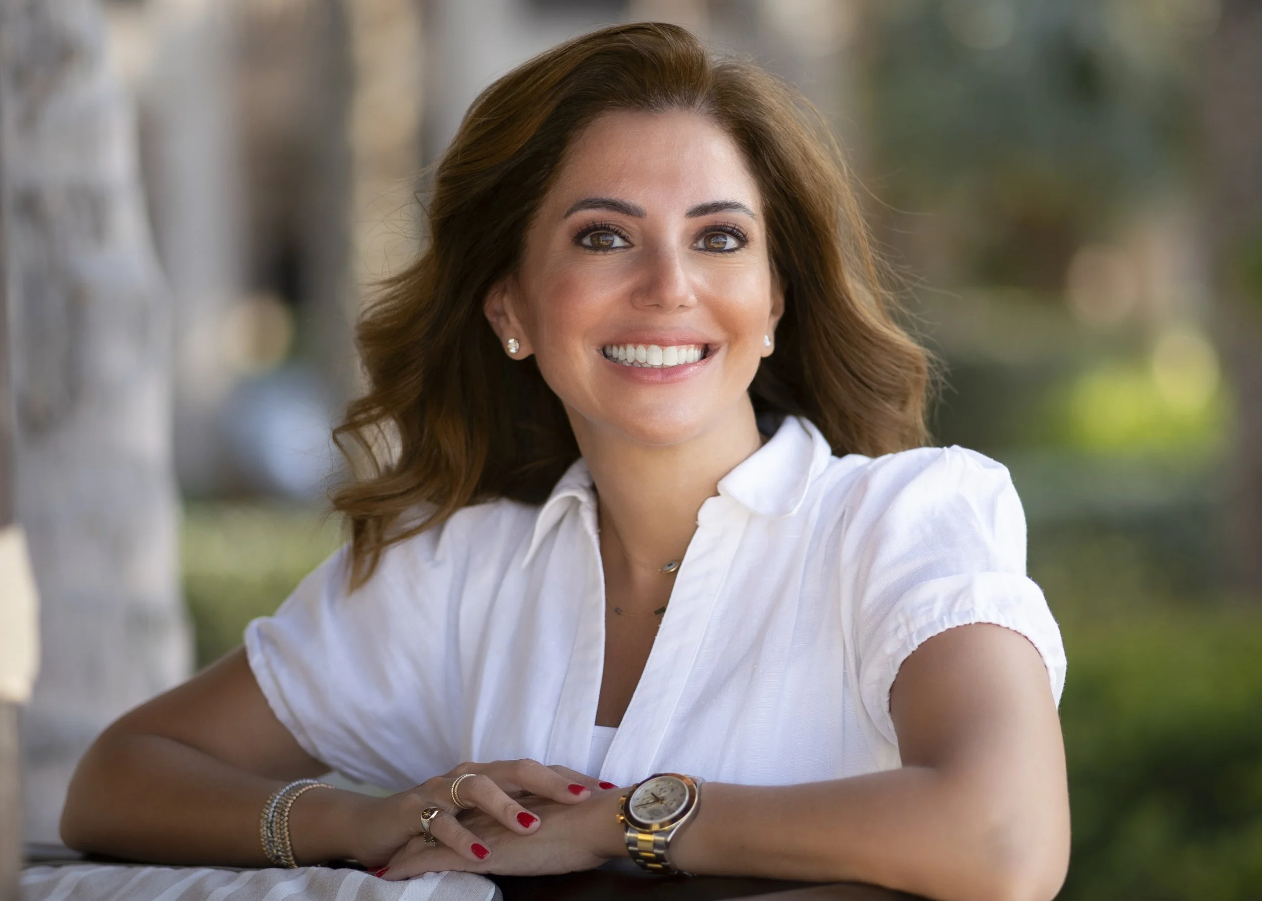 A woman with brown hair, smiling, wearing a white shirt, and a watch, sitting outdoors with a blurred background.