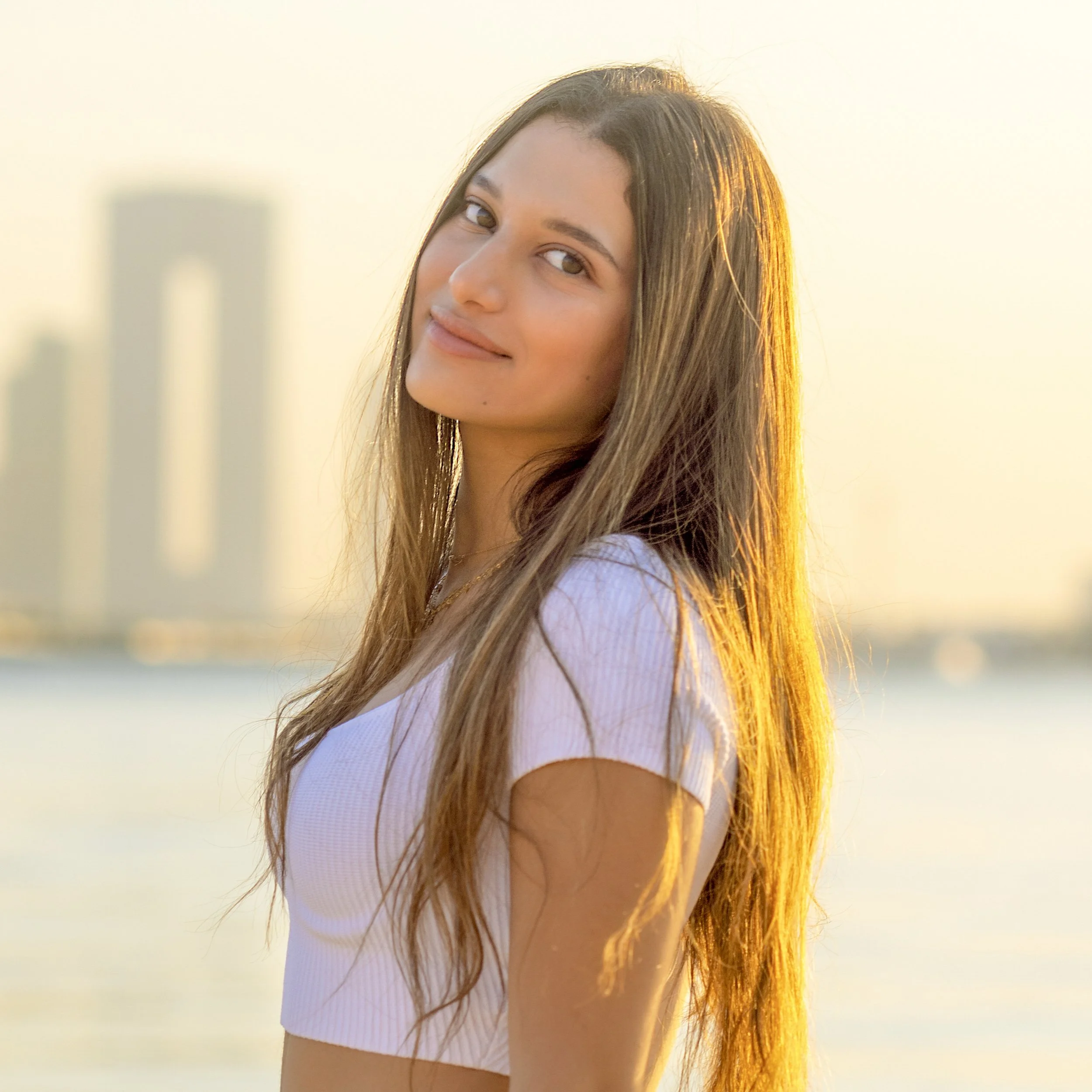 Young woman with long hair looking at camera during sunset by the water with city skyline in the background.