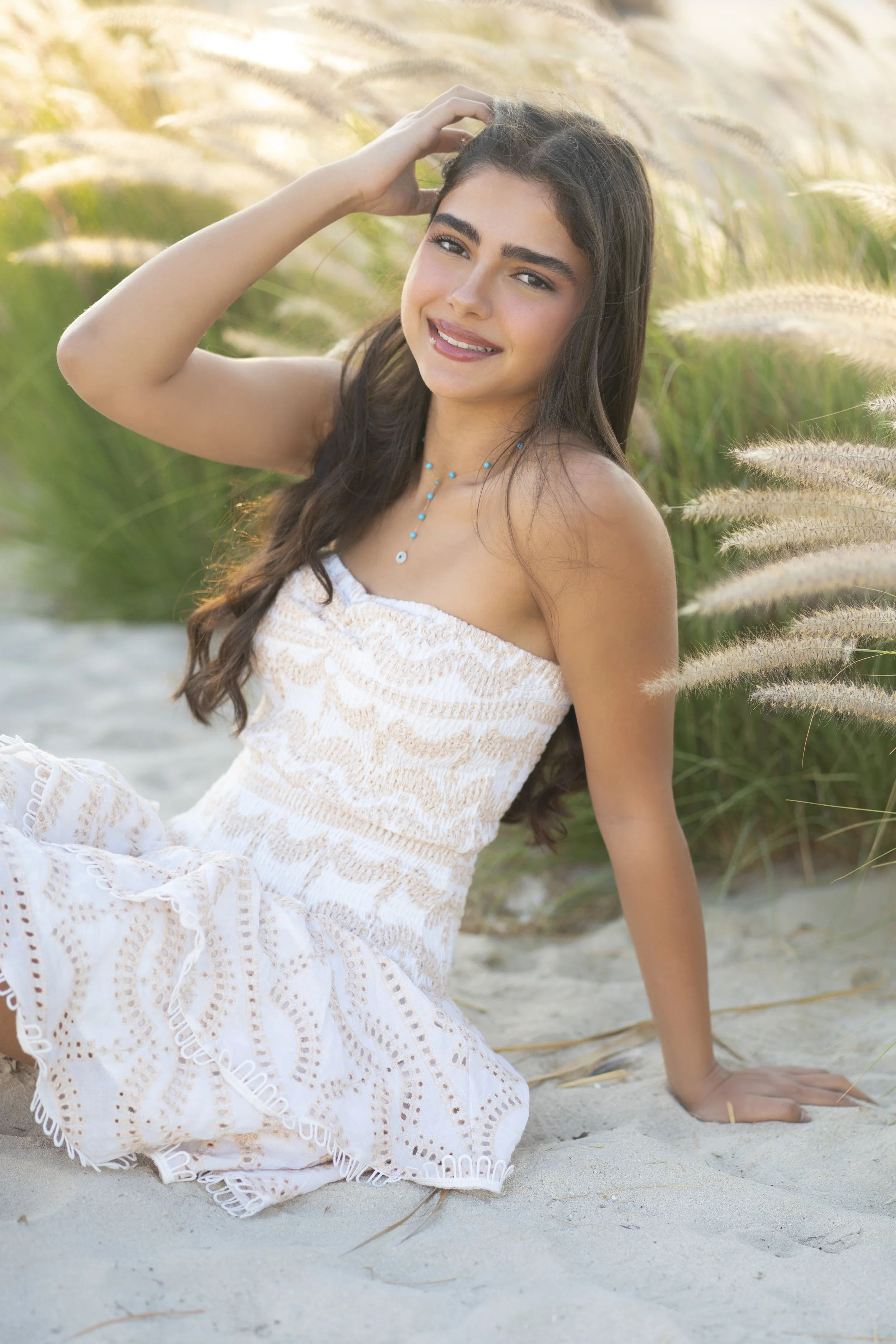 A young woman sitting on sandy beach in front of tall grass, smiling and posing for the camera. She has long, dark hair and is wearing a white strapless dress and turquoise jewelry.