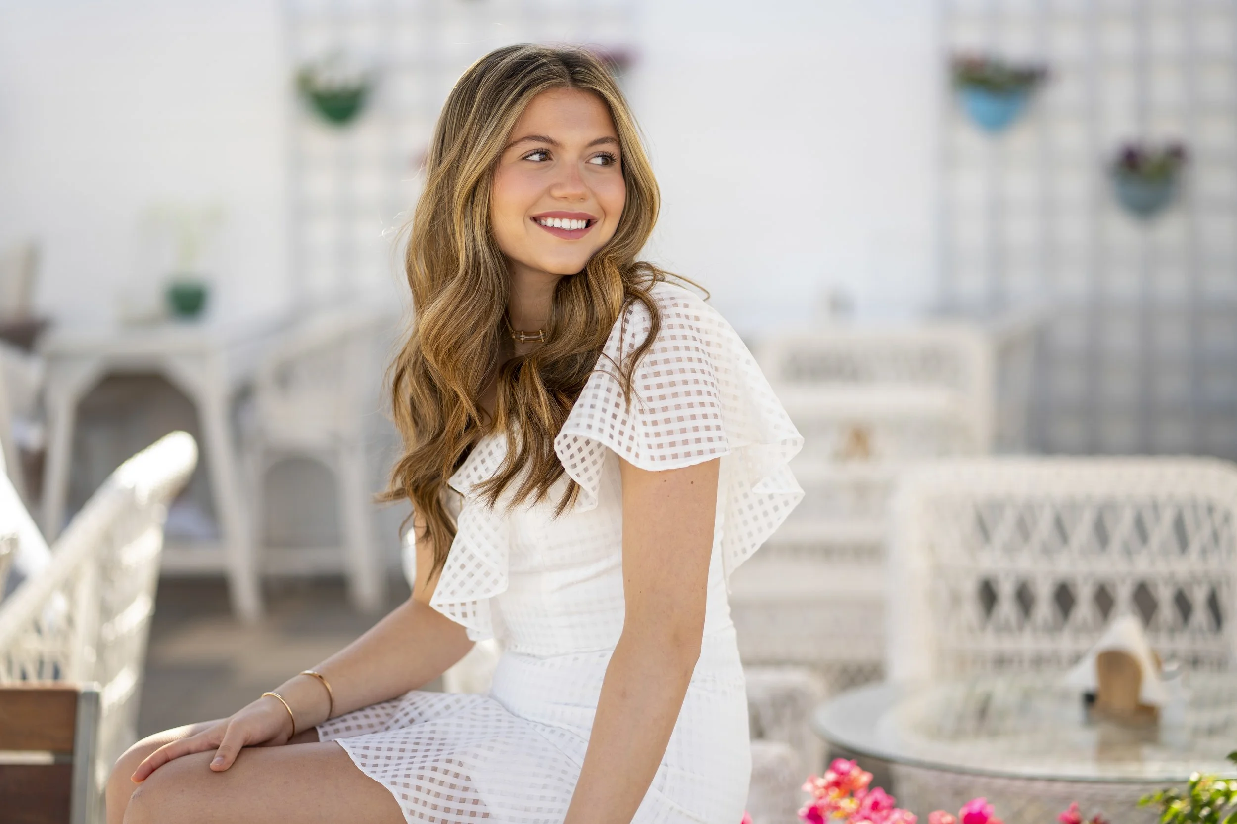 A young woman with long, wavy blonde hair smiling and looking to her left, sitting outdoors in a bright, airy space with white wicker furniture and colorful plants.