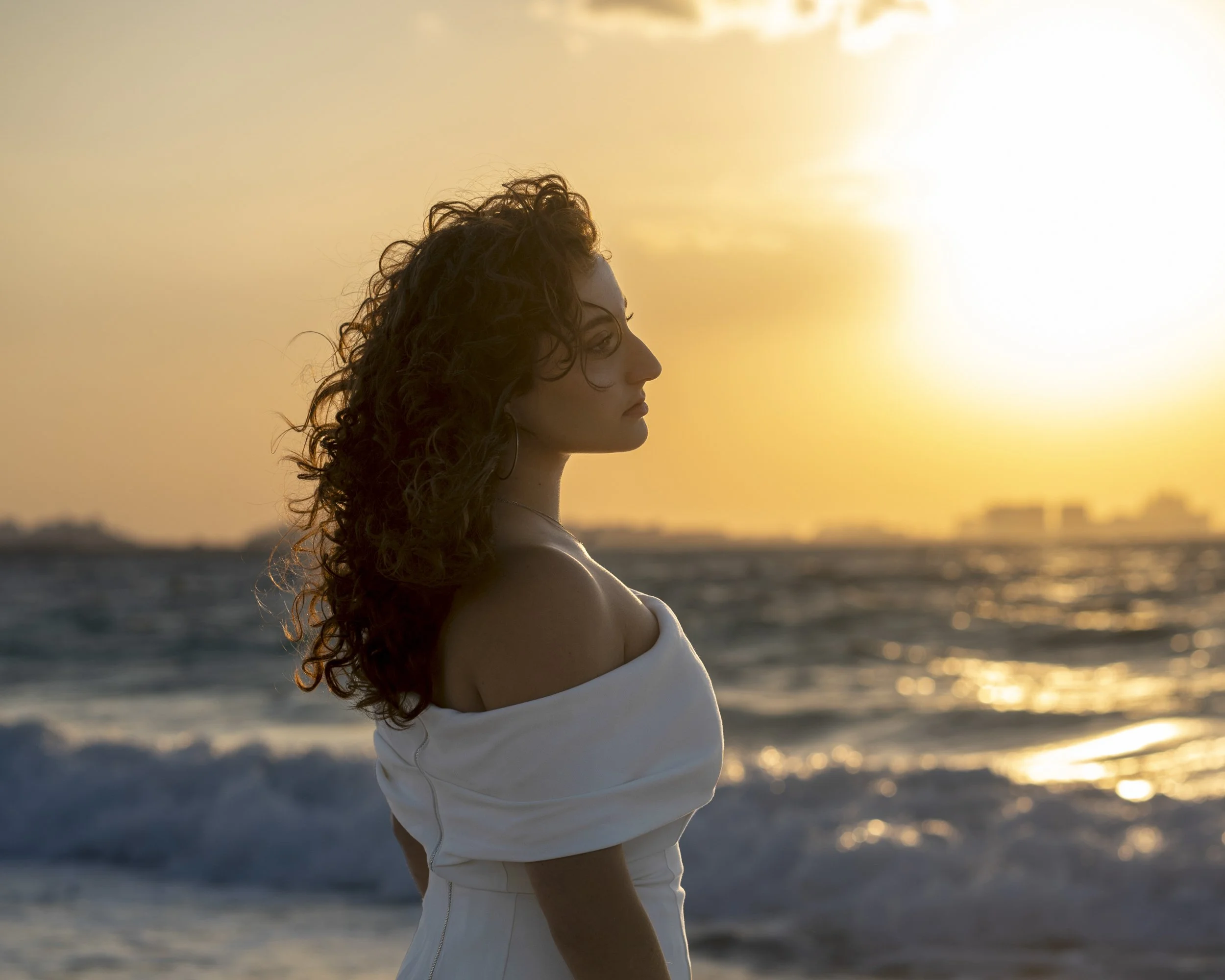 A woman with curly hair standing on a beach at sunset, wearing a white off-shoulder dress.