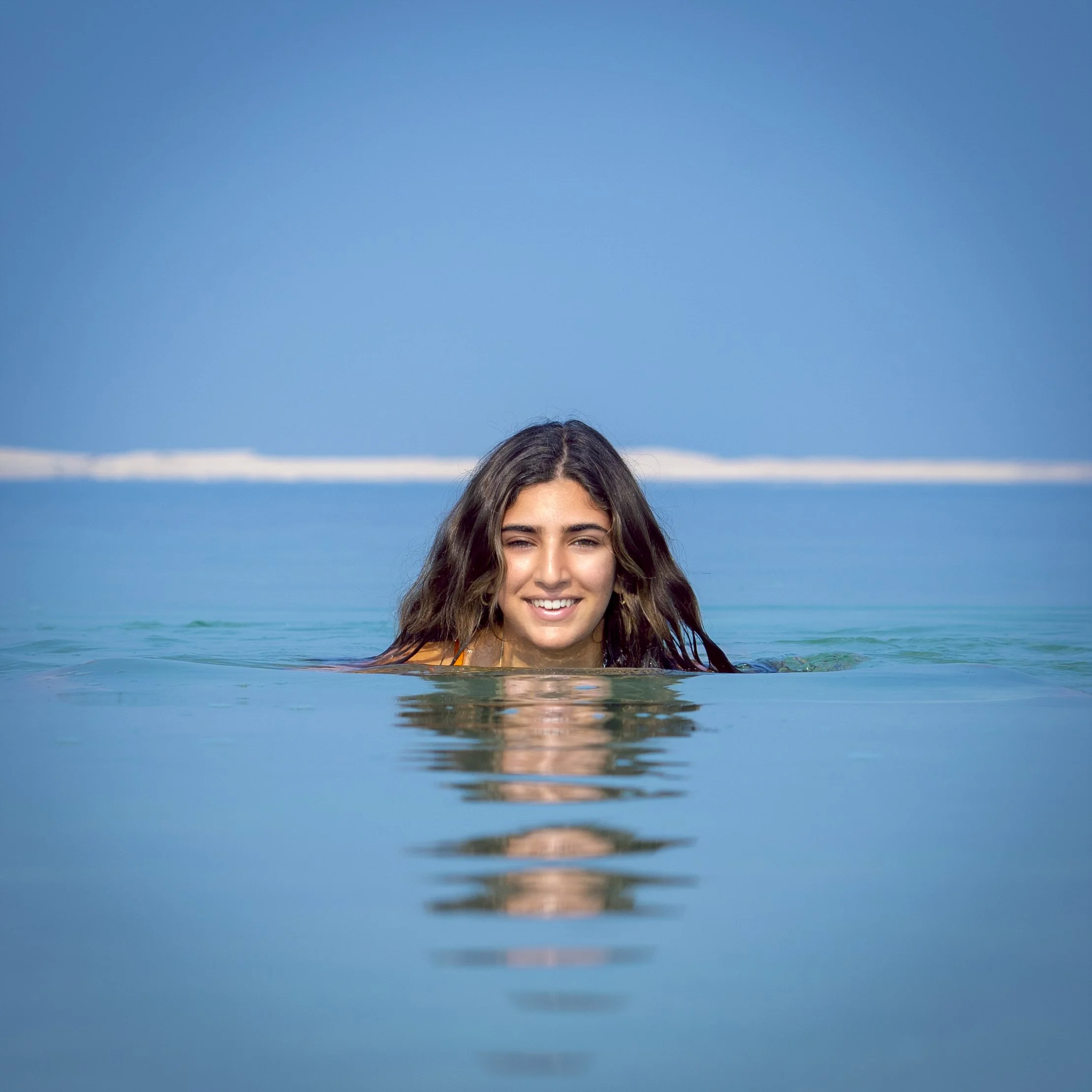 A young woman with long, wavy brown hair swimming in the ocean with a blue sky overhead.