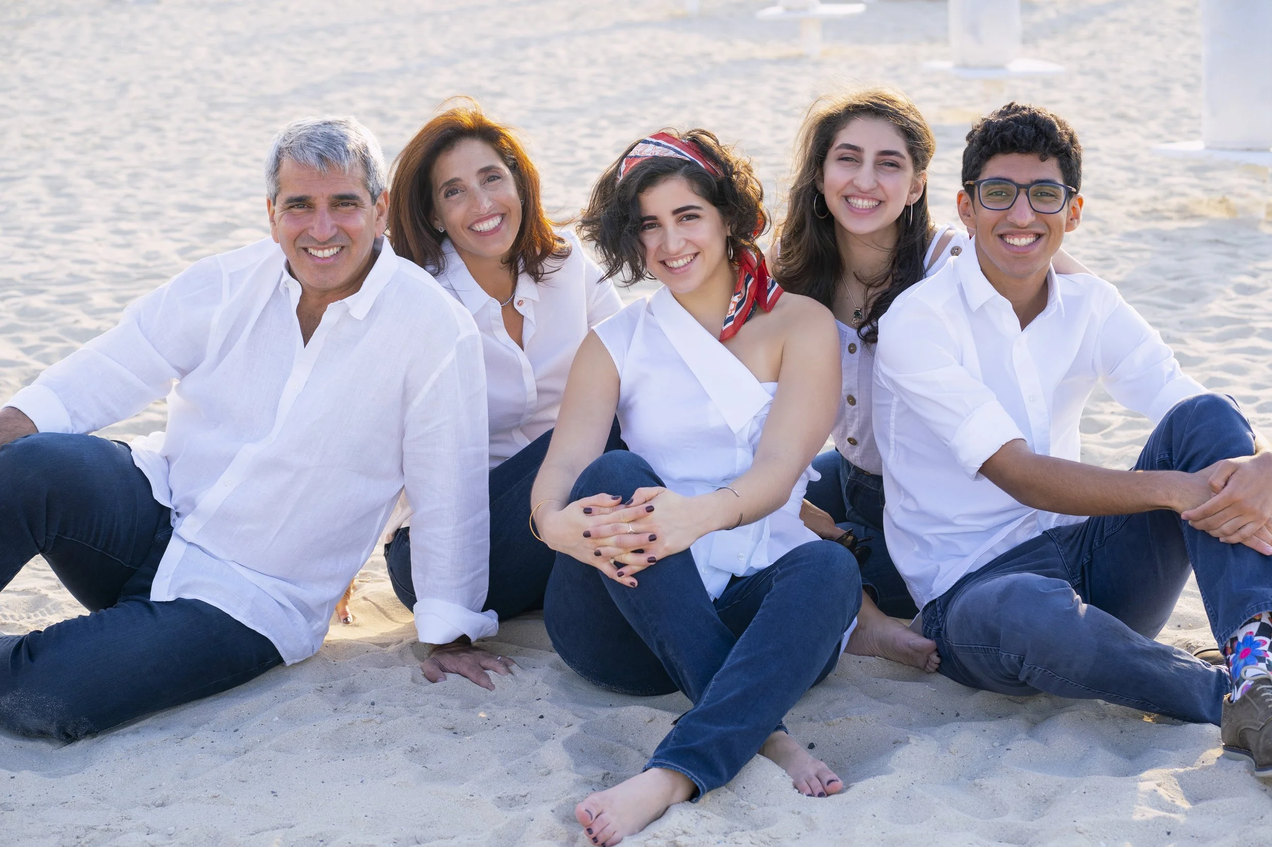 A family of six sitting on the beach during daytime, smiling. They are dressed in casual white shirts and blue jeans, with some wearing accessories like a bandana and earrings. The background shows sandy beach and water.