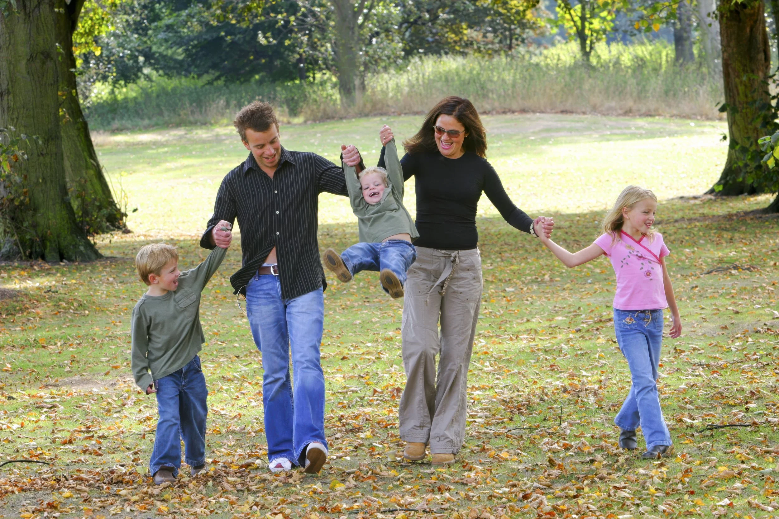 A family of five, including two adults and three children, is playing outdoors in a park during autumn. The adults are holding a young boy and girl’s hands, swinging them in the air as they walk through fallen leaves. Everyone is smiling and enjoying