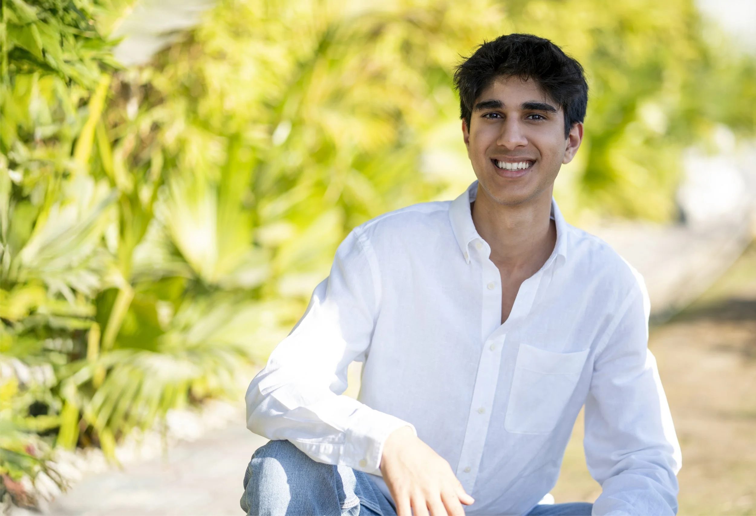 A young man with black hair and a bright smile, wearing a white shirt, outdoors on a sunny day surrounded by green foliage.