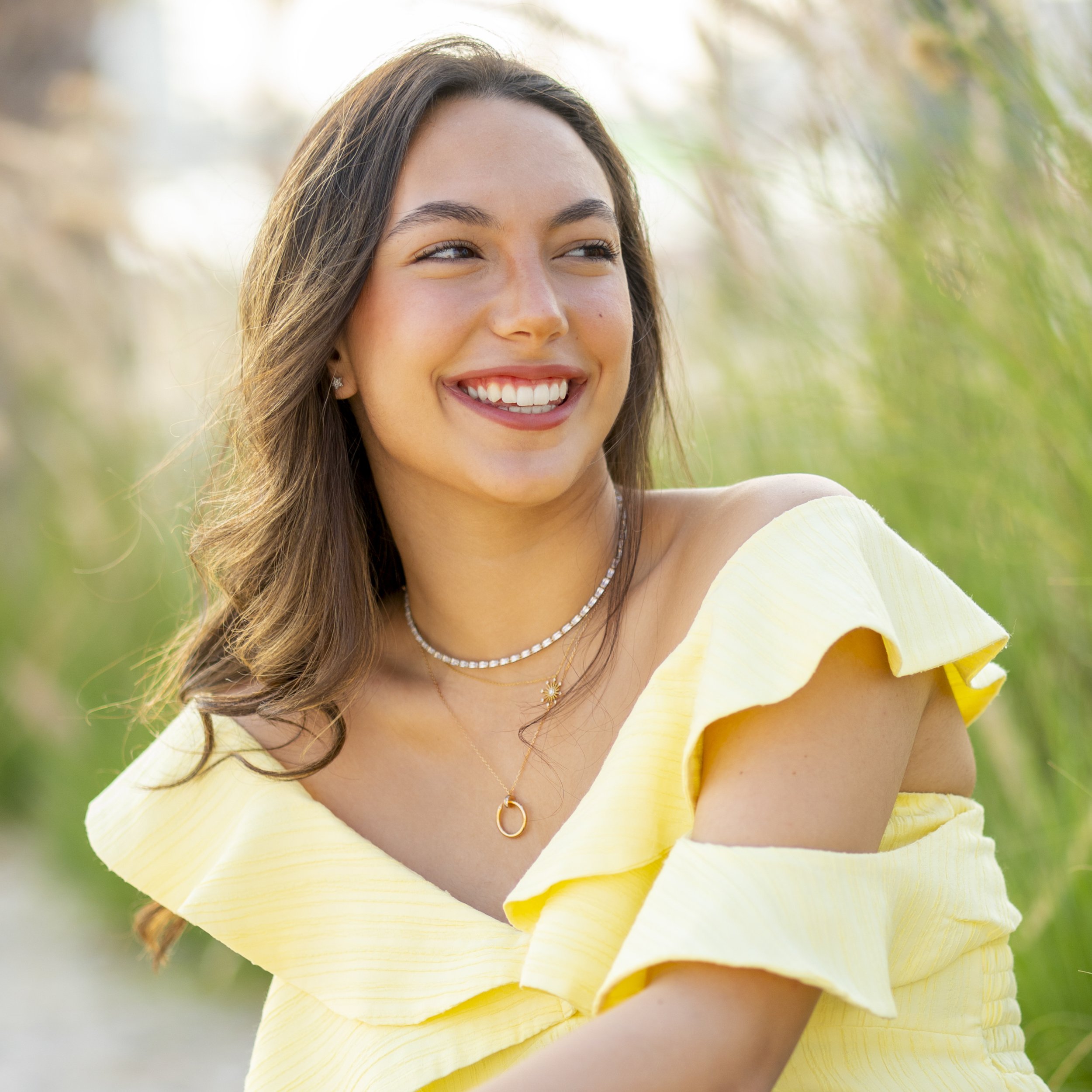 A young woman with long brown hair, smiling with white teeth, wearing a yellow off-shoulder dress and layered necklaces, standing outdoors with green blurred grass background.