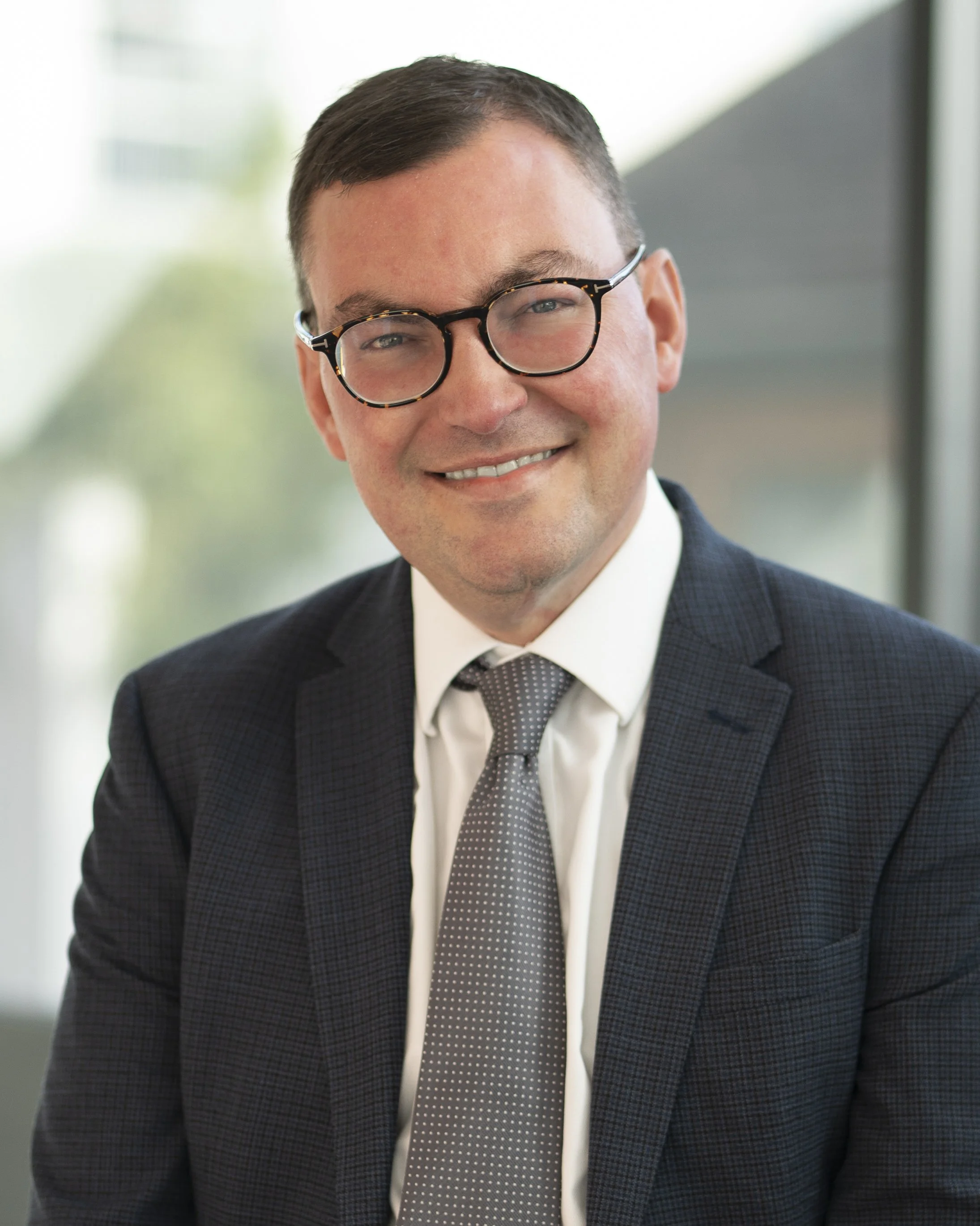 A smiling man with glasses in a business suit and tie in a professional office setting.