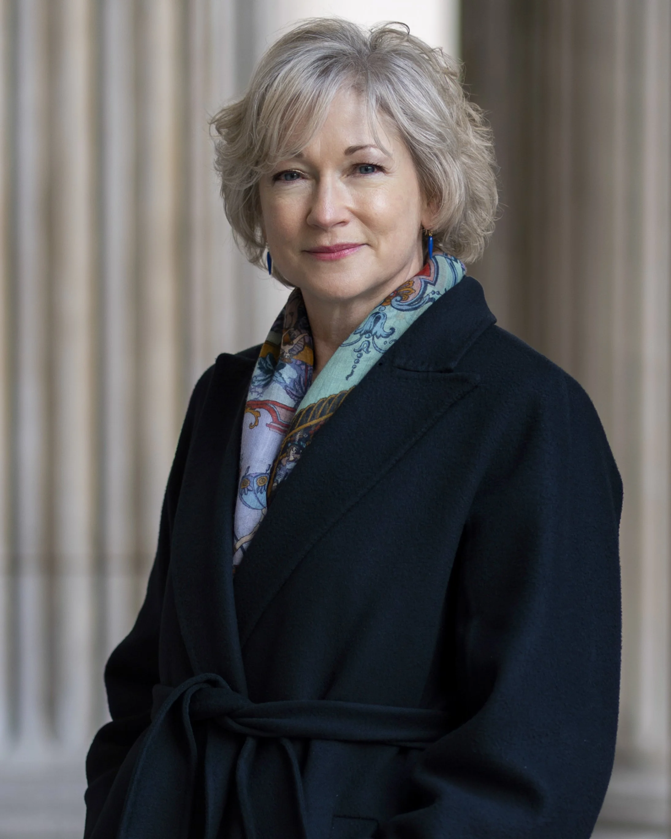 A middle-aged woman with short blonde hair wearing a dark coat and colorful scarf, standing in front of a background with columns.