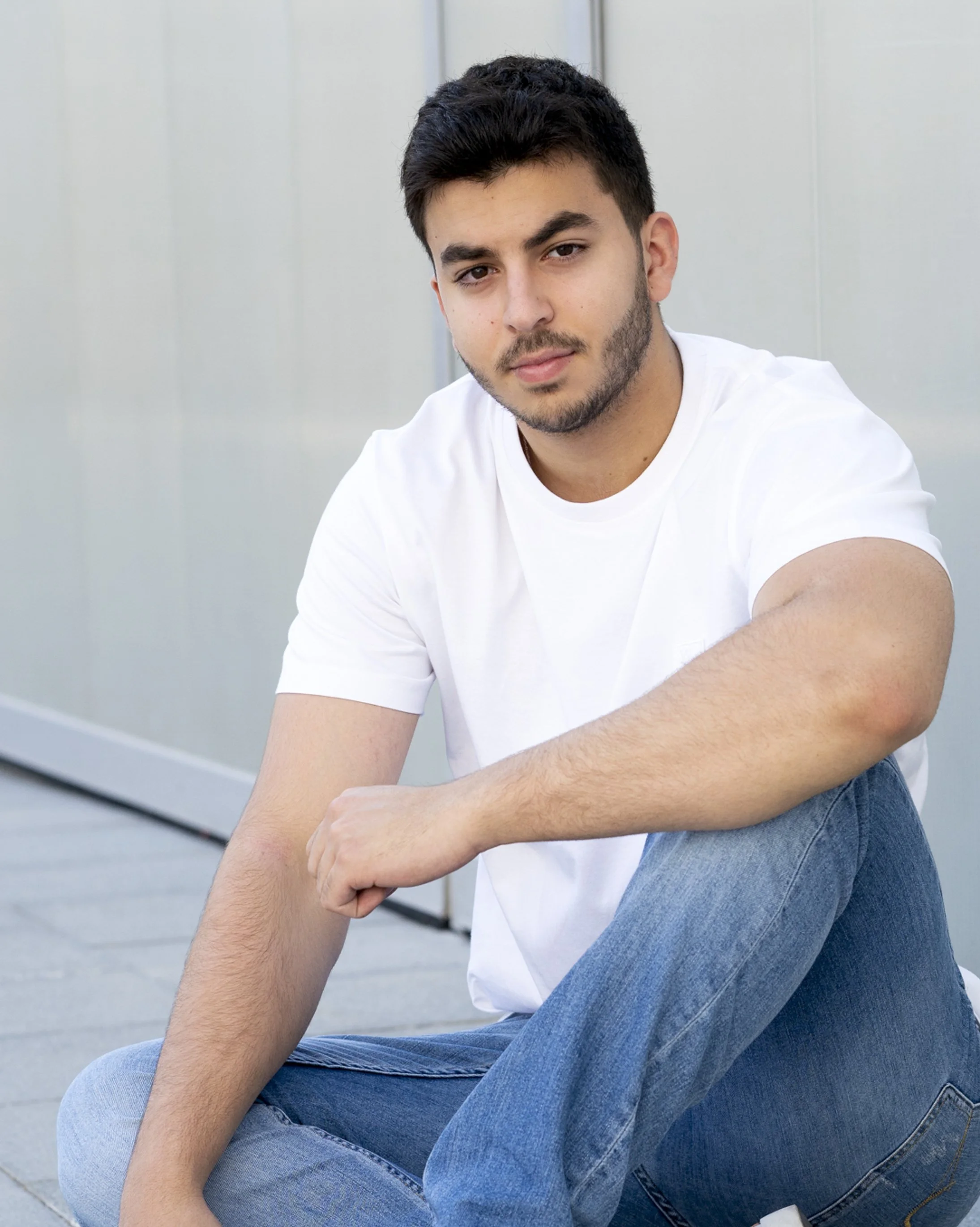 A young man with dark hair and facial hair, sitting outdoors against a modern building wall, wearing a white t-shirt and blue jeans, looking into the camera with a relaxed expression.