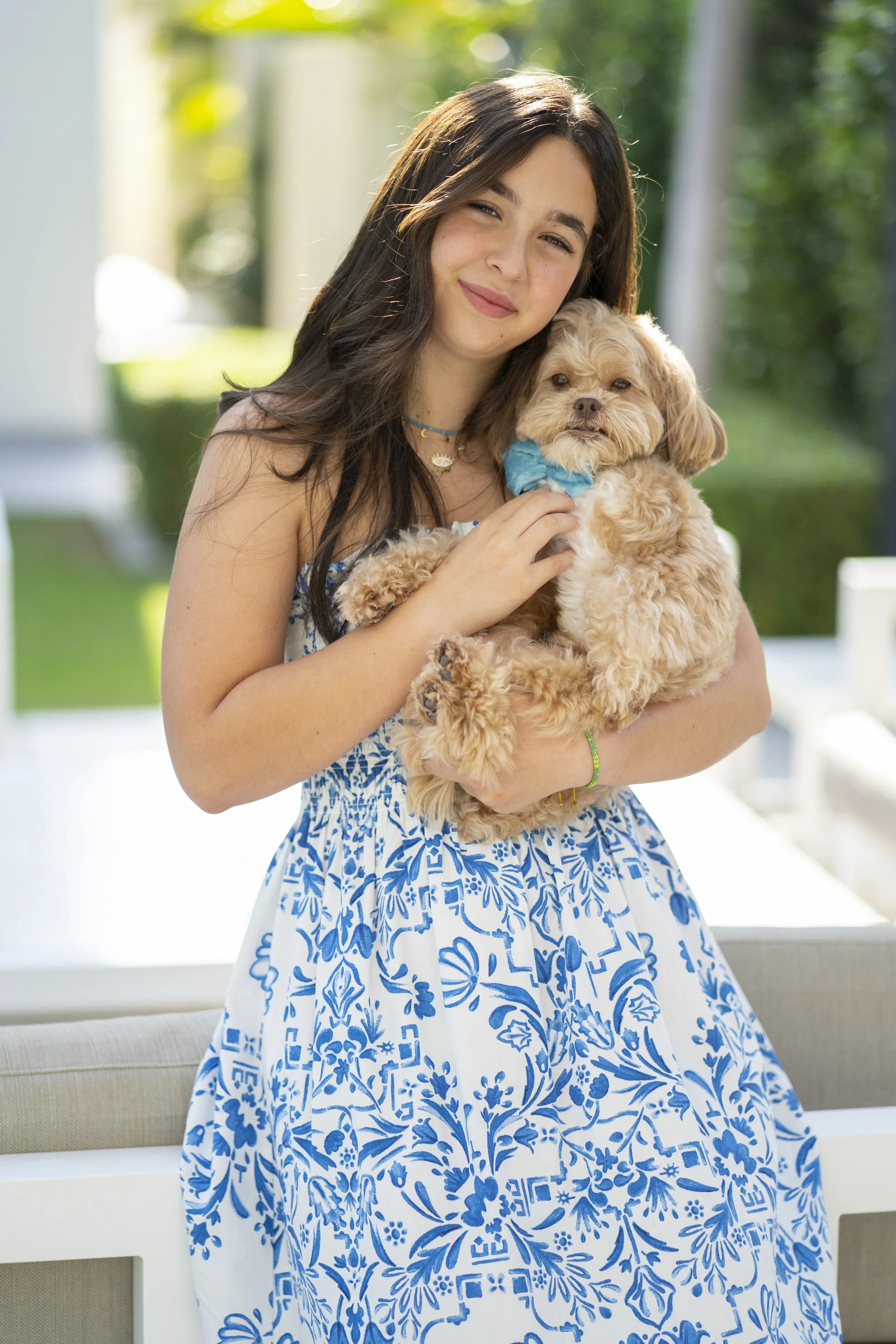 A young woman with long dark hair, wearing a white and blue patterned dress, holding a small fluffy beige dog with a blue collar outdoors on a sunny day.