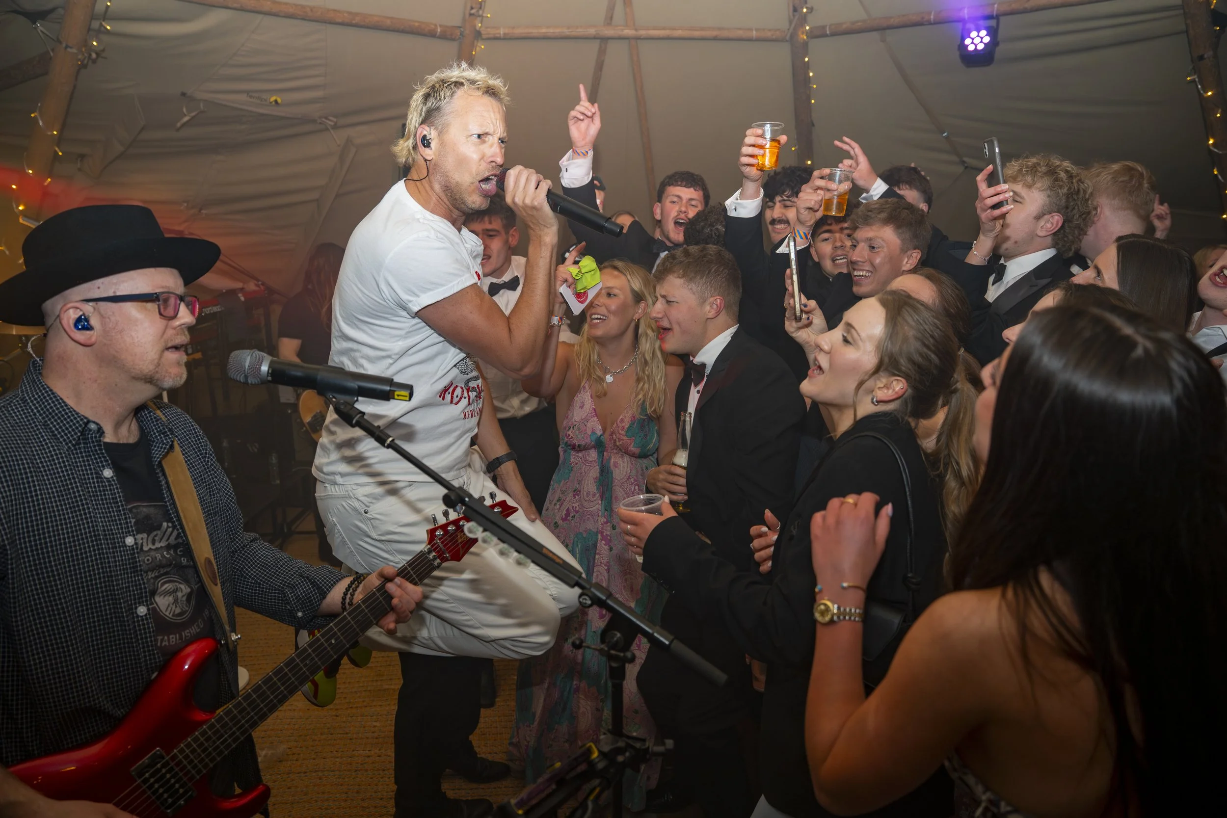 A lively party scene with a singer performing with a microphone, a guitarist, and a crowd of young adults enjoying the music and celebrating under a tent decorated with string lights.