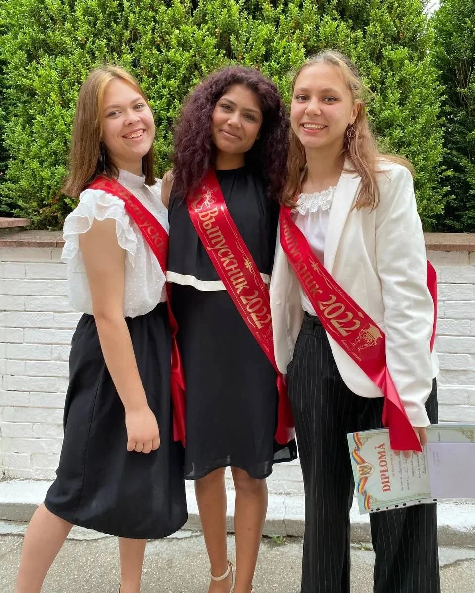 Three young women standing side by side outdoors, wearing graduation sashes for Bujumbura 2022, smiling, with a white brick wall and green shrubbery in the background.