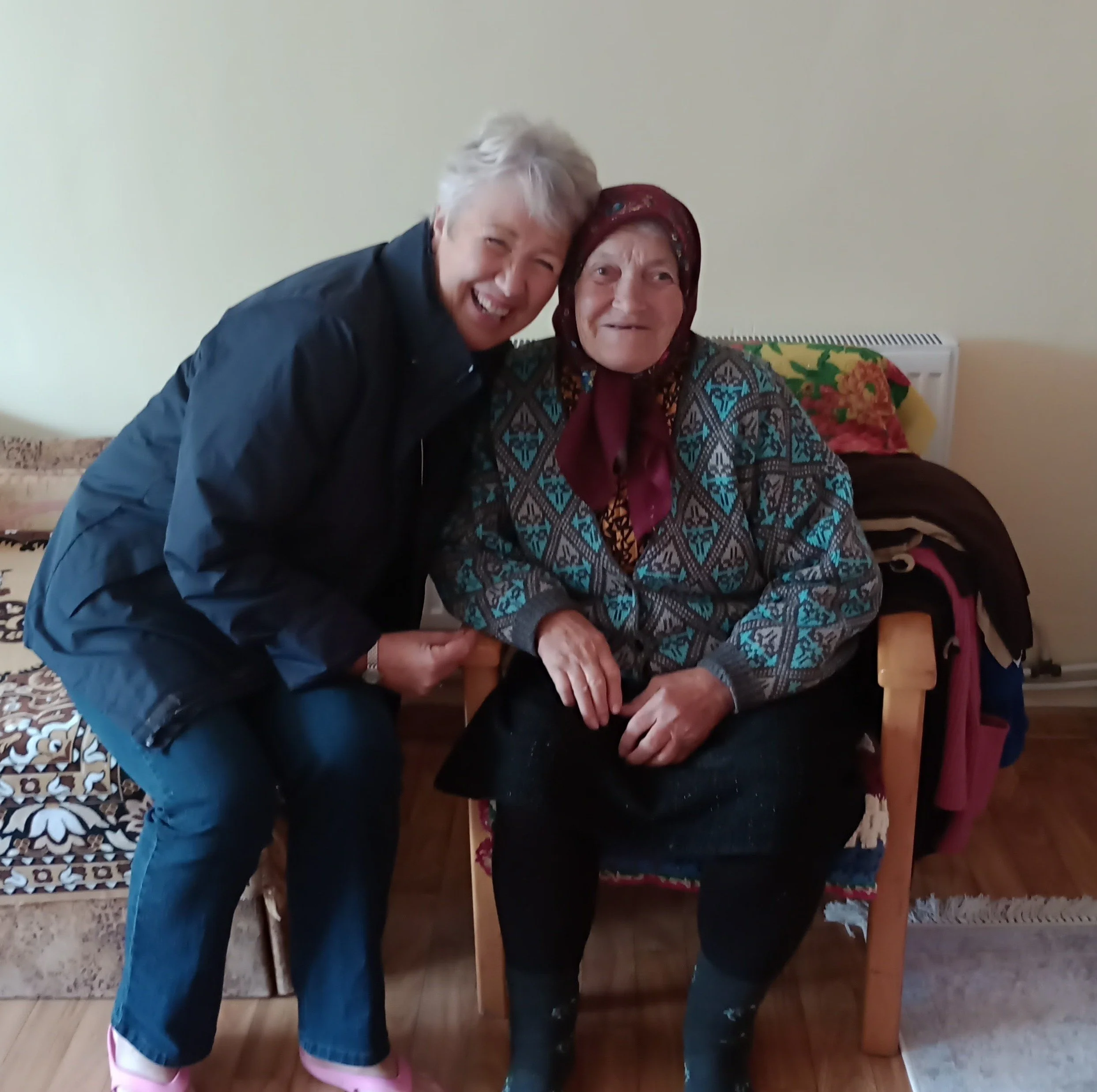 A young man in a black jacket and blue jeans hugging an elderly woman seated in a chair, both smiling, in a cozy room with a sofa and blankets in the background.