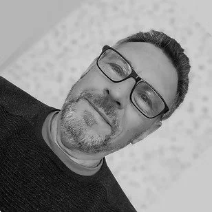 A man with glasses and facial hair taking a selfie in front of a light background.