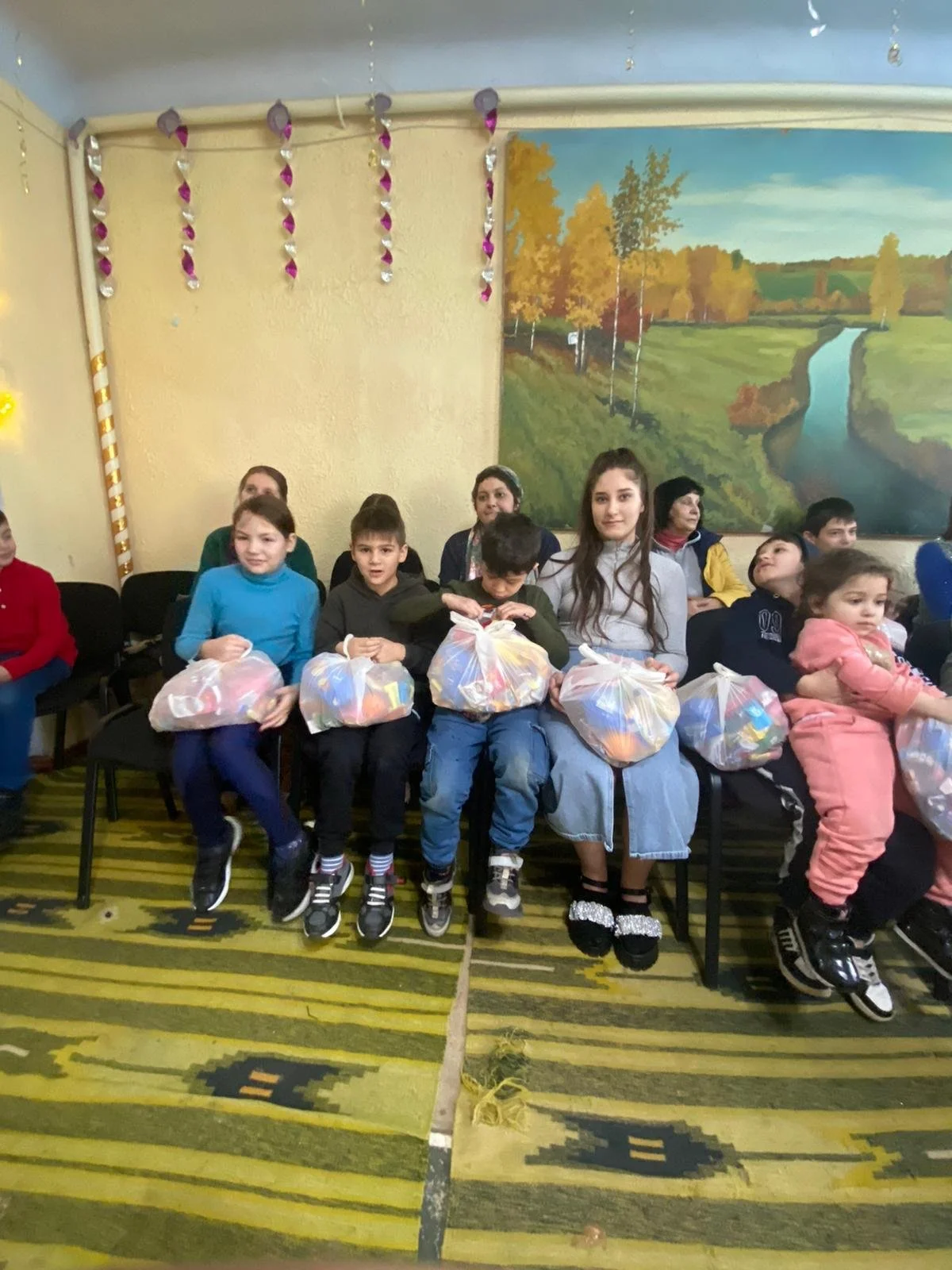 Children sitting in a row holding plastic bags filled with snacks or gifts in a room decorated for a celebration or event.