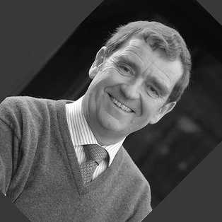Black and white portrait of a smiling man in a sweater and collared shirt, against a dark background.
