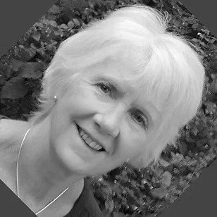 Black and white photo of a smiling woman with short, light-colored hair outdoors, with foliage in the background.