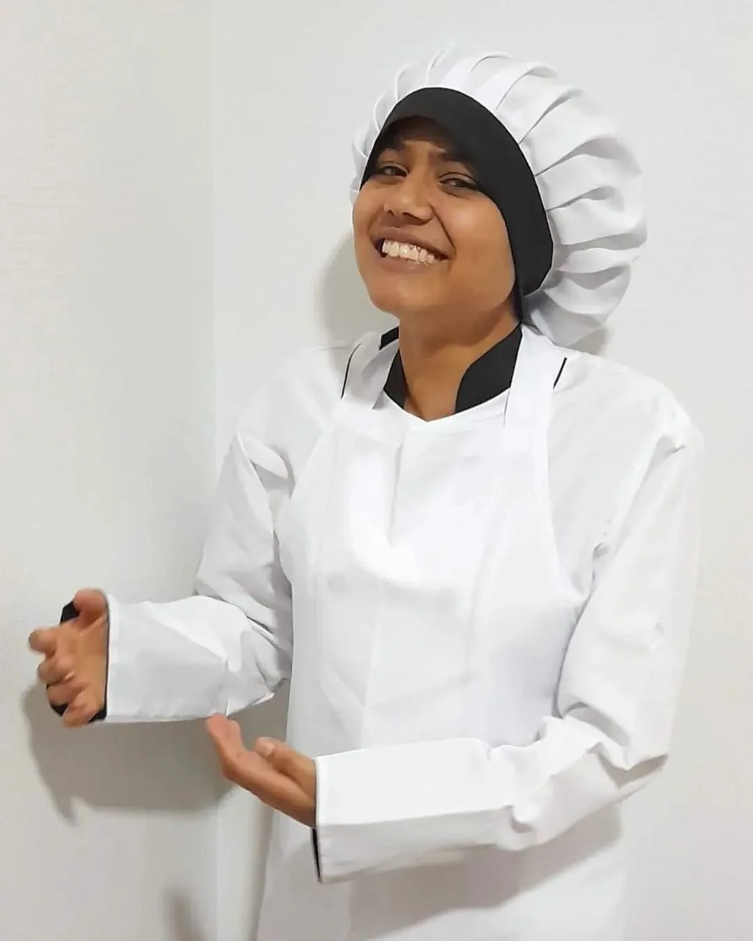Person wearing a chef's hat and chef's coat, smiling, standing against a plain white wall.