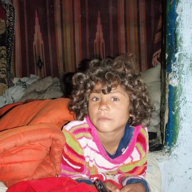 A young child with curly hair, wearing a colorful striped sweater, sitting on a bed with an orange blanket, looking at the camera with a serious expression.