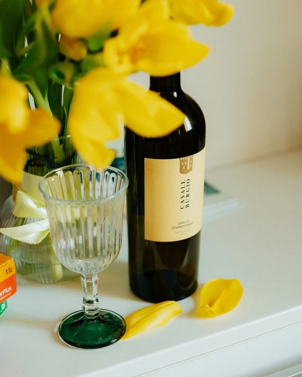A table with a bottle of Casale Buracío Chardonnay wine, a decorative glass, yellow flowers with some petals on the table, and a small box in the background.