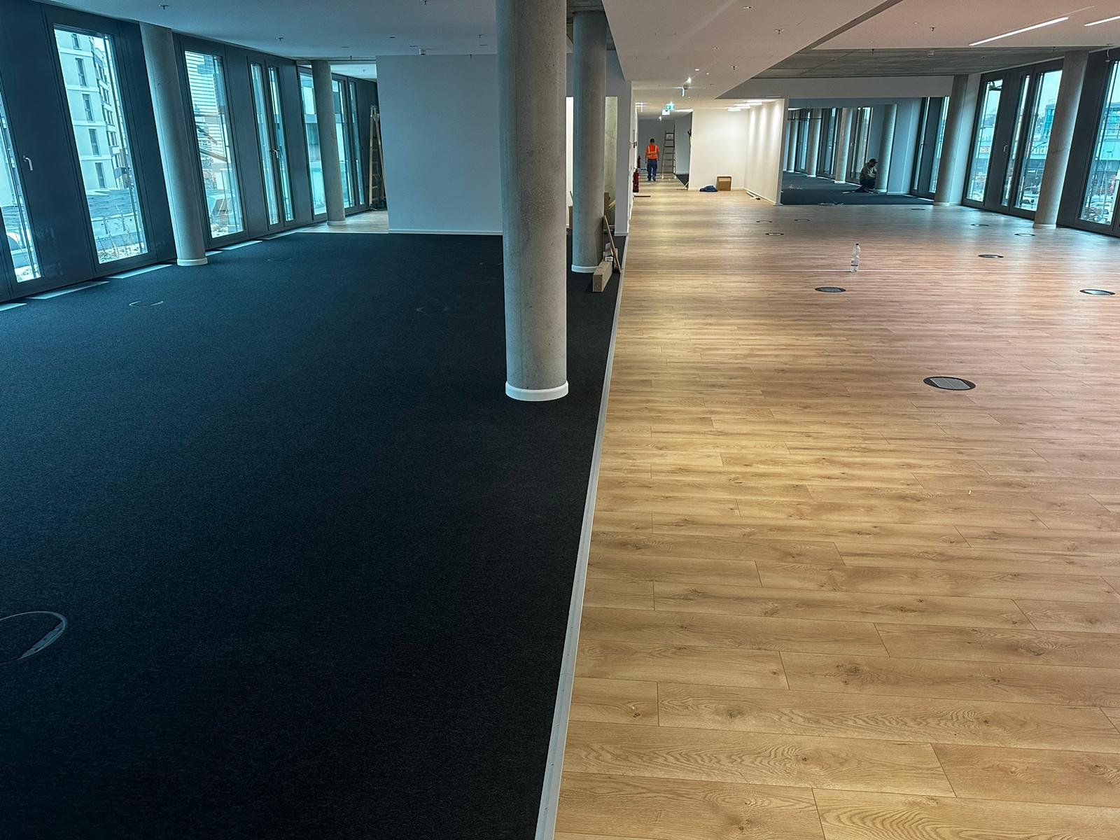 Innenraum eines Gebäudes mit großen Fenstern, zwei unterschiedlich große Bodenbeläge (schwarzer Teppich und Holzdielen), einige Baustellenutensilien und Personen im Hintergrund.