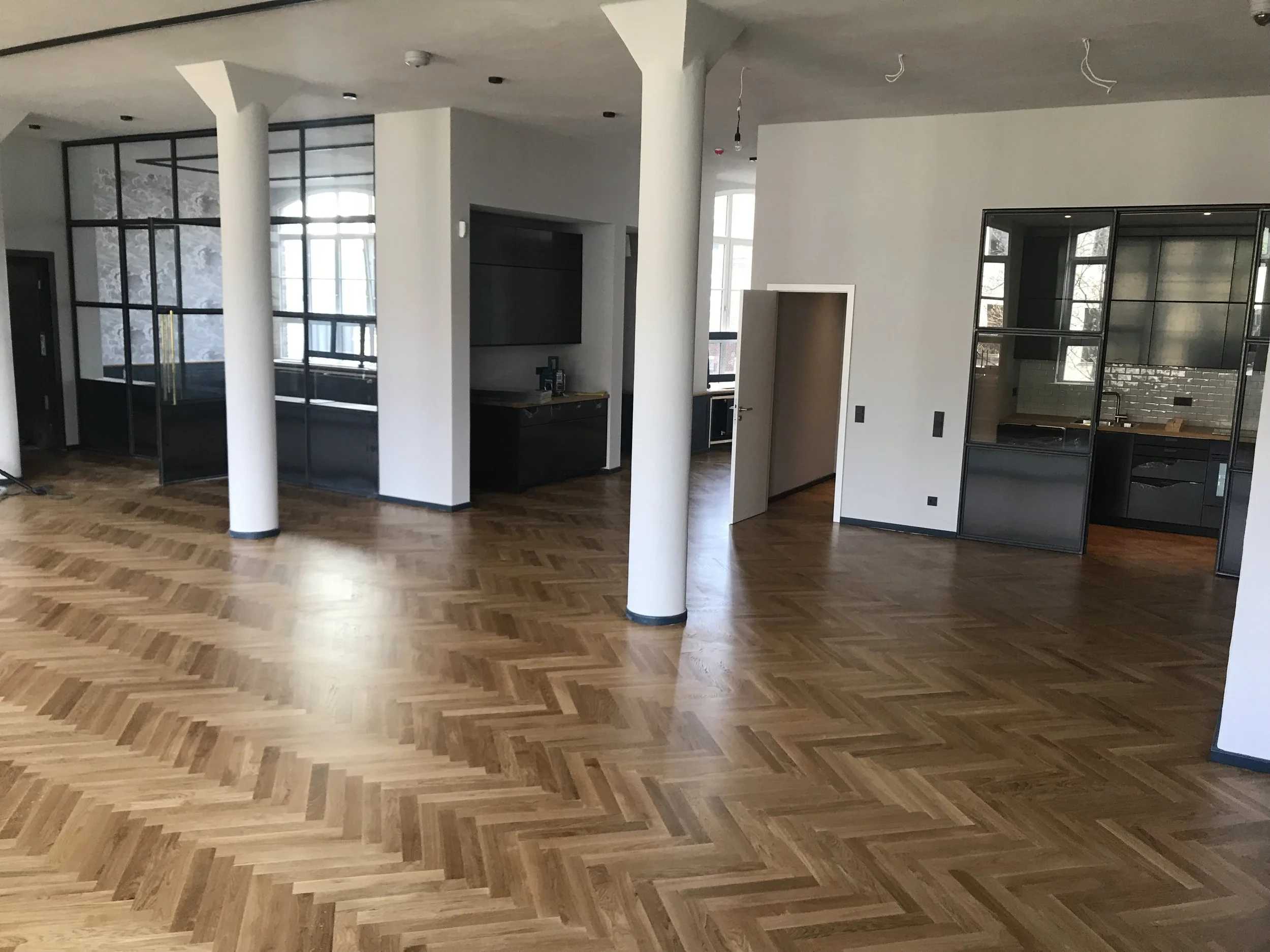 Innenansicht eines noch unfertigen Hauses mit großen Fenstern, Holzboden in Fischgrätmuster, weißen Wänden, schwarzen Küchen- und Wohnzimmerschränken und sichtbaren Kabeln an der Decke.